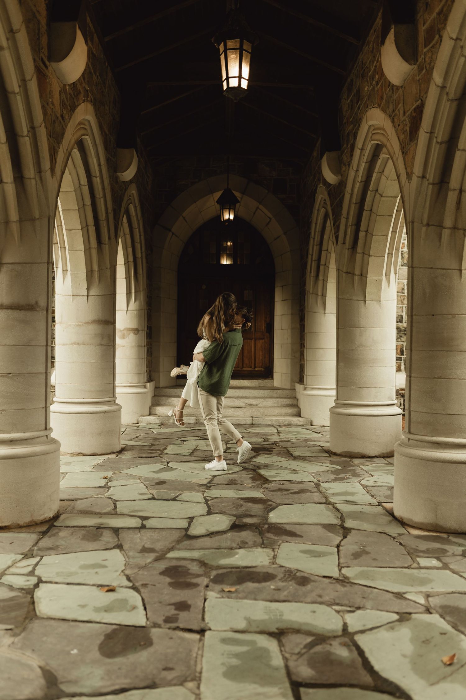 Gothic stone archways and columns line a dimly lit corridor with rough stone flooring and hanging lanterns.