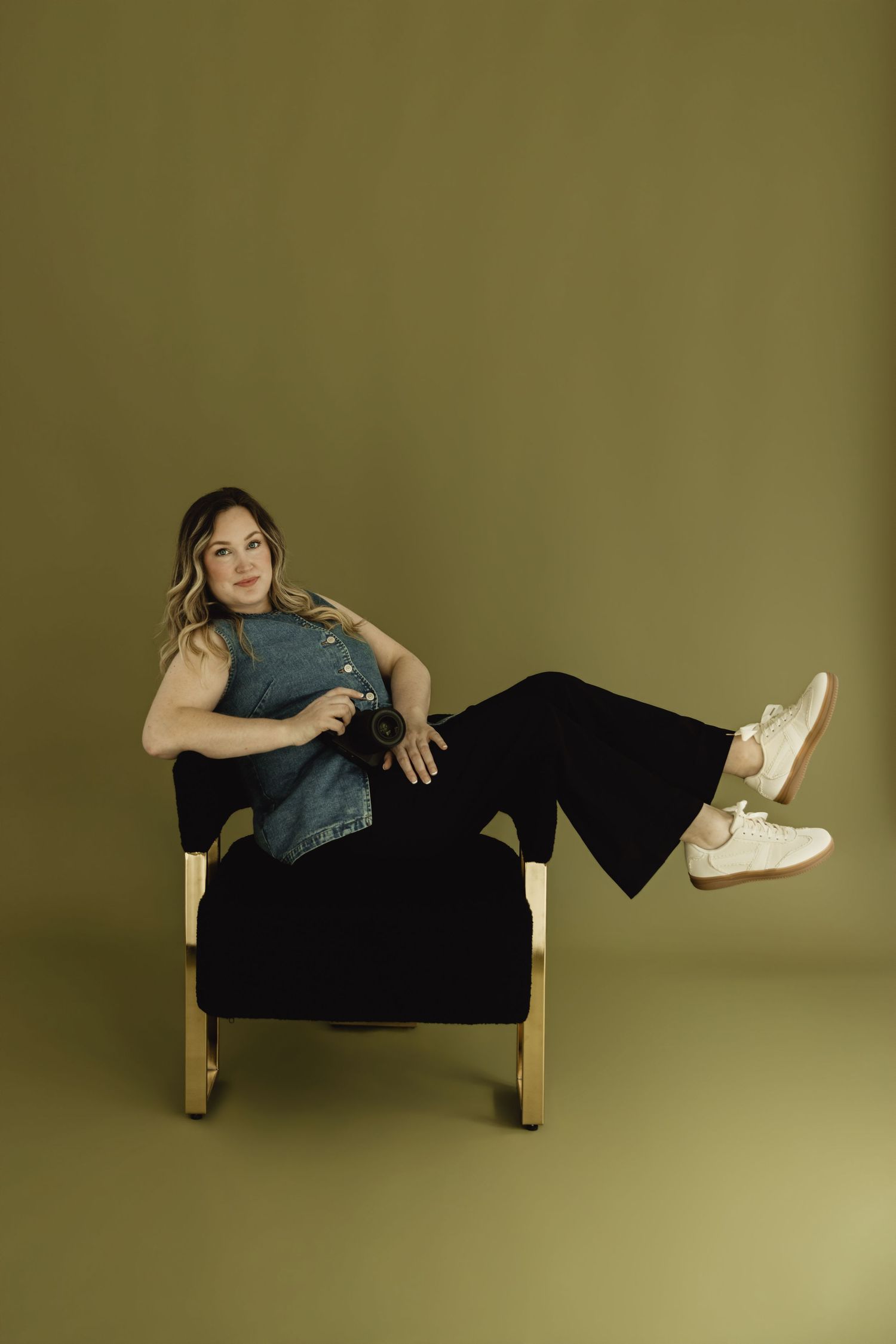 Woman in denim vest and black wide-leg pants sits in a chair holding a camera against an olive green background.