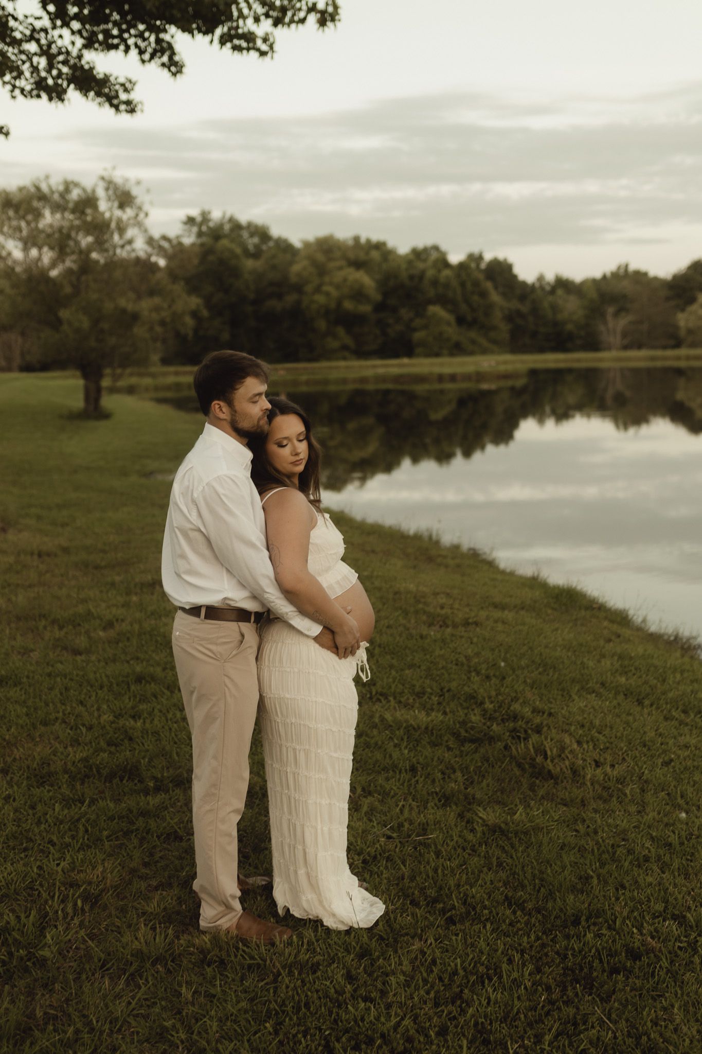 Couple poses by a serene lake at dusk, man embraces pregnant woman in white dress on lush green grass.
