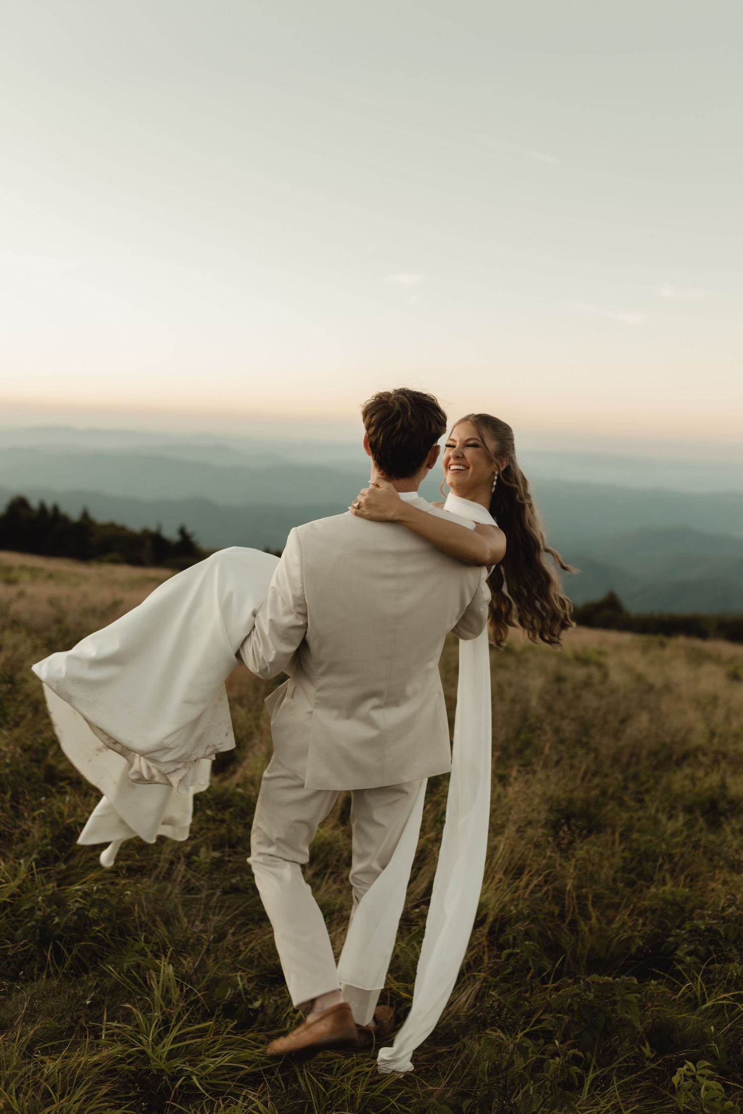 Groom carrying bride in white dress on mountain meadow at sunset with scenic misty hills in background.