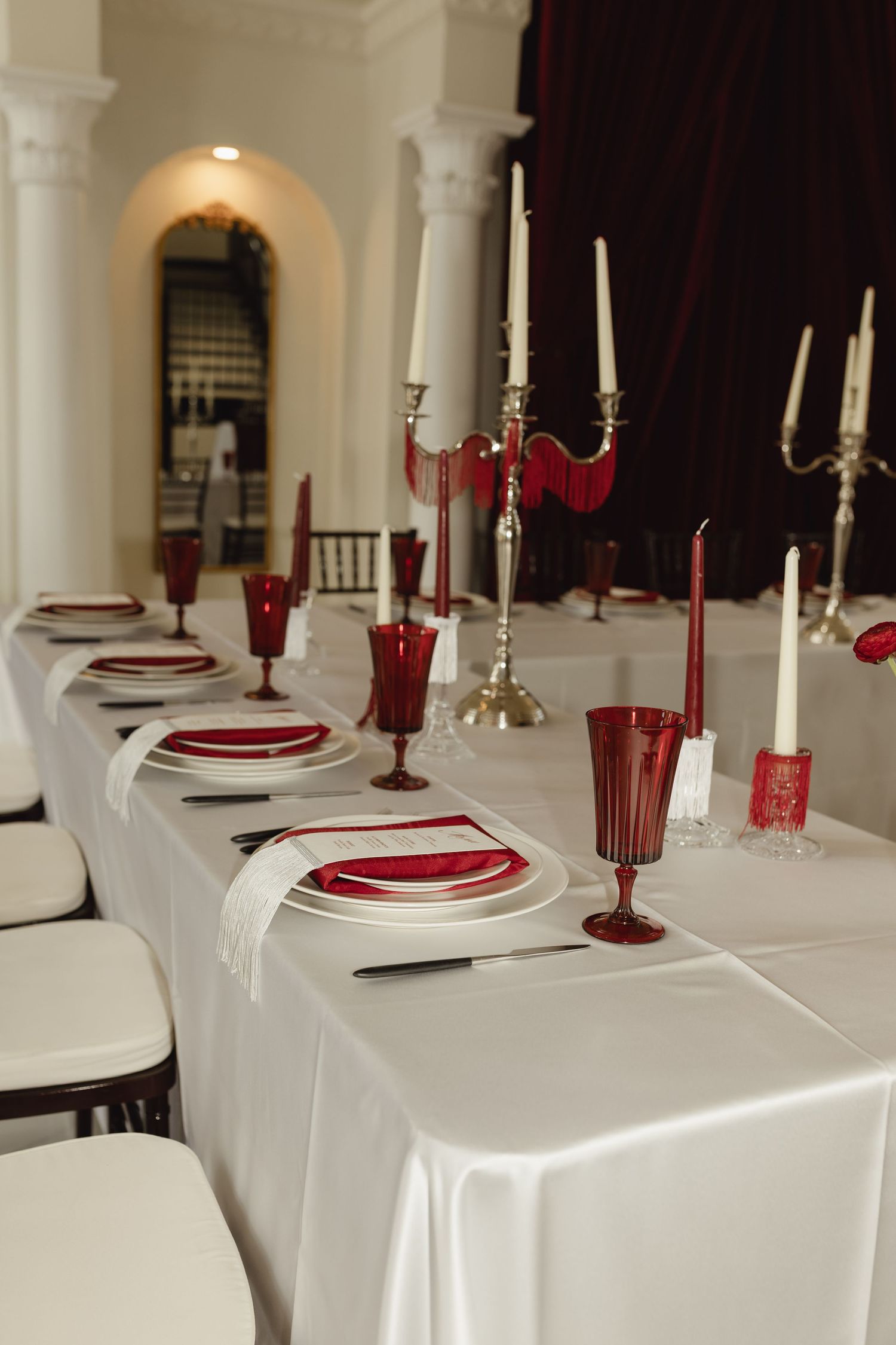 Elegant red and white wedding table setting with candelabras, crimson glassware, and white plates on a satin tablecloth.