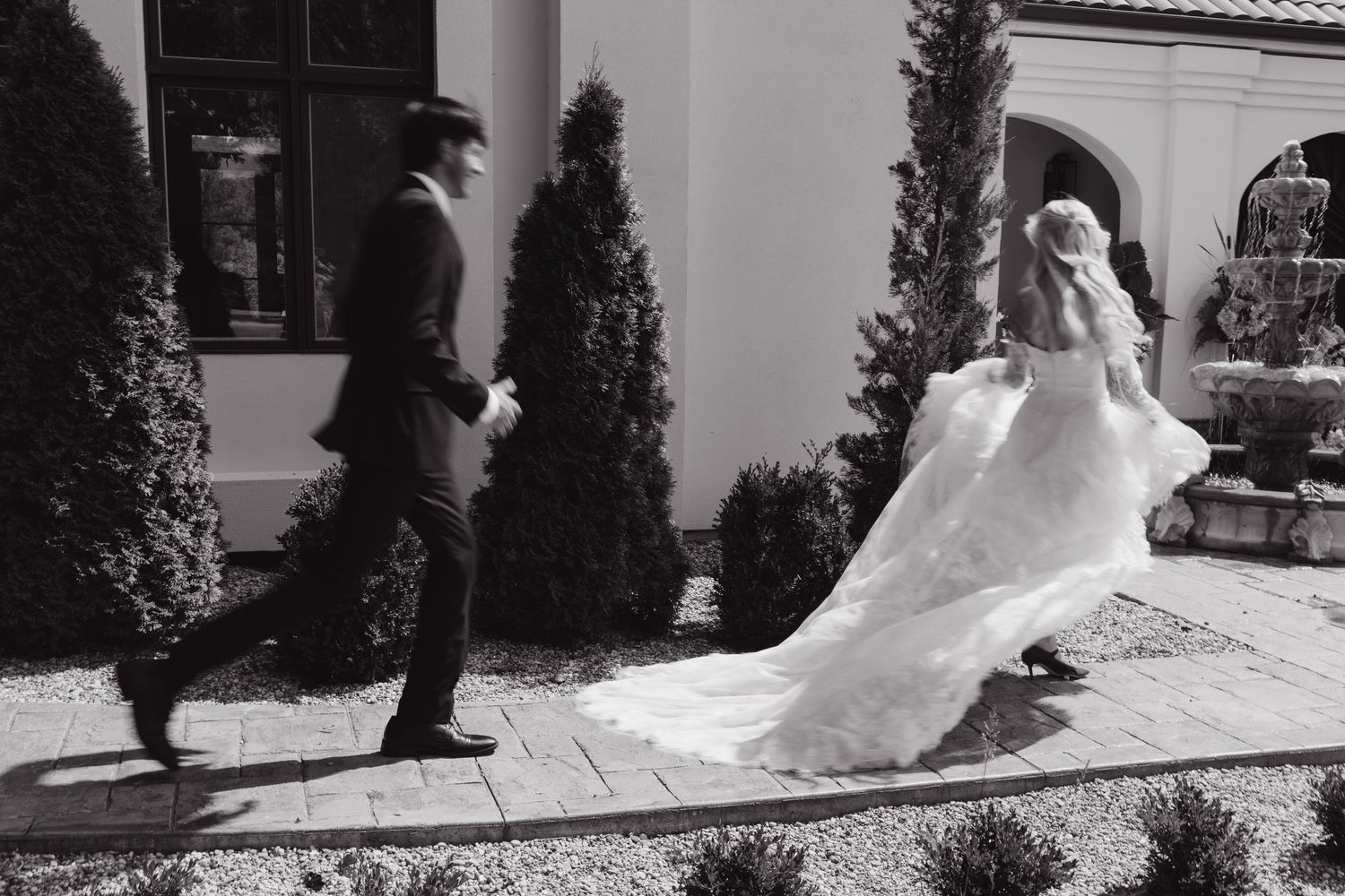 Black and white photo of a bride in a flowing gown and groom walking together outside a elegant venue.