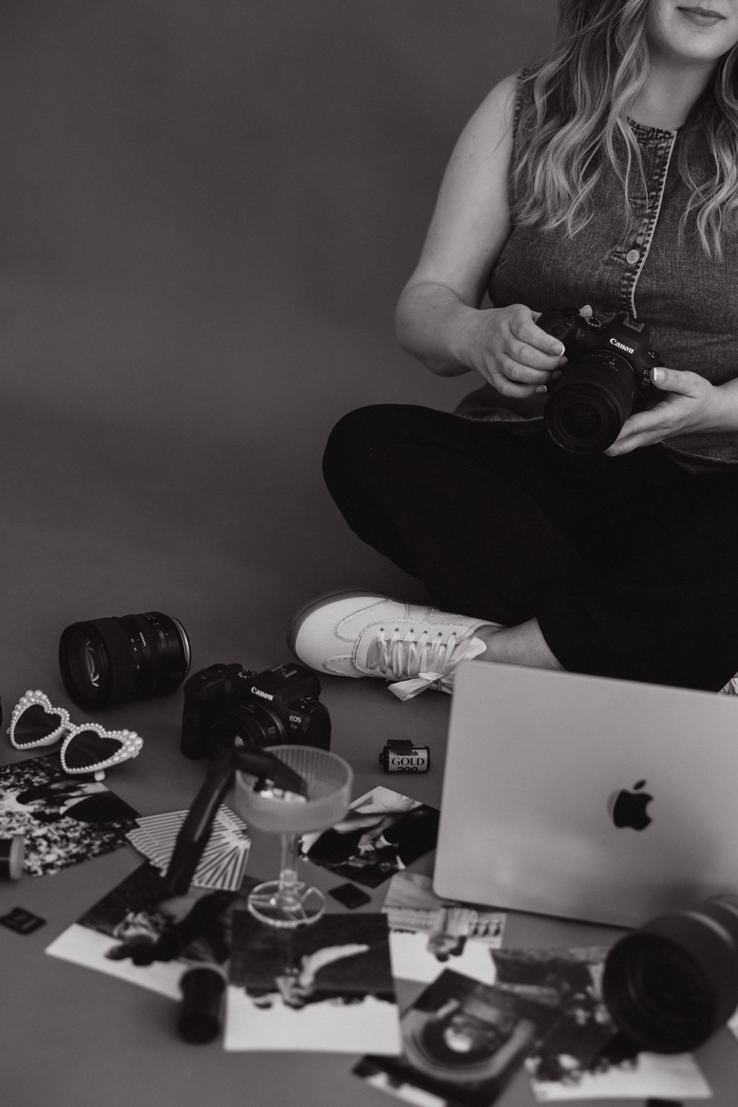 Black and white photo of a woman sitting cross-legged holding a camera surrounded by photography gear and an Apple laptop.