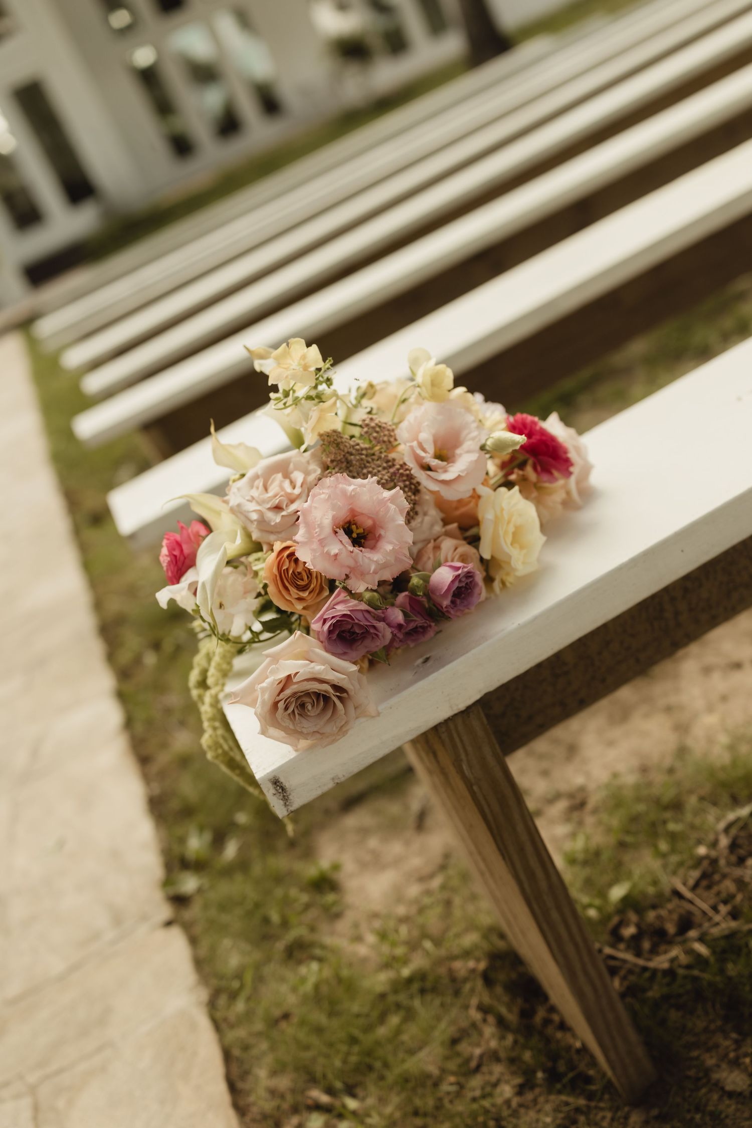 Floral bouquet with pink roses and purple flowers resting on a white wooden bench at an outdoor wedding ceremony.
