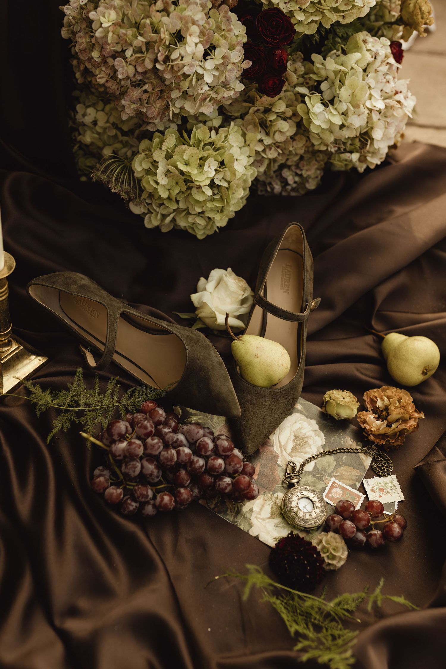 Elegant still life with vintage shoes, grapes, pears, pocket watch, and hydrangeas on brown silk fabric.