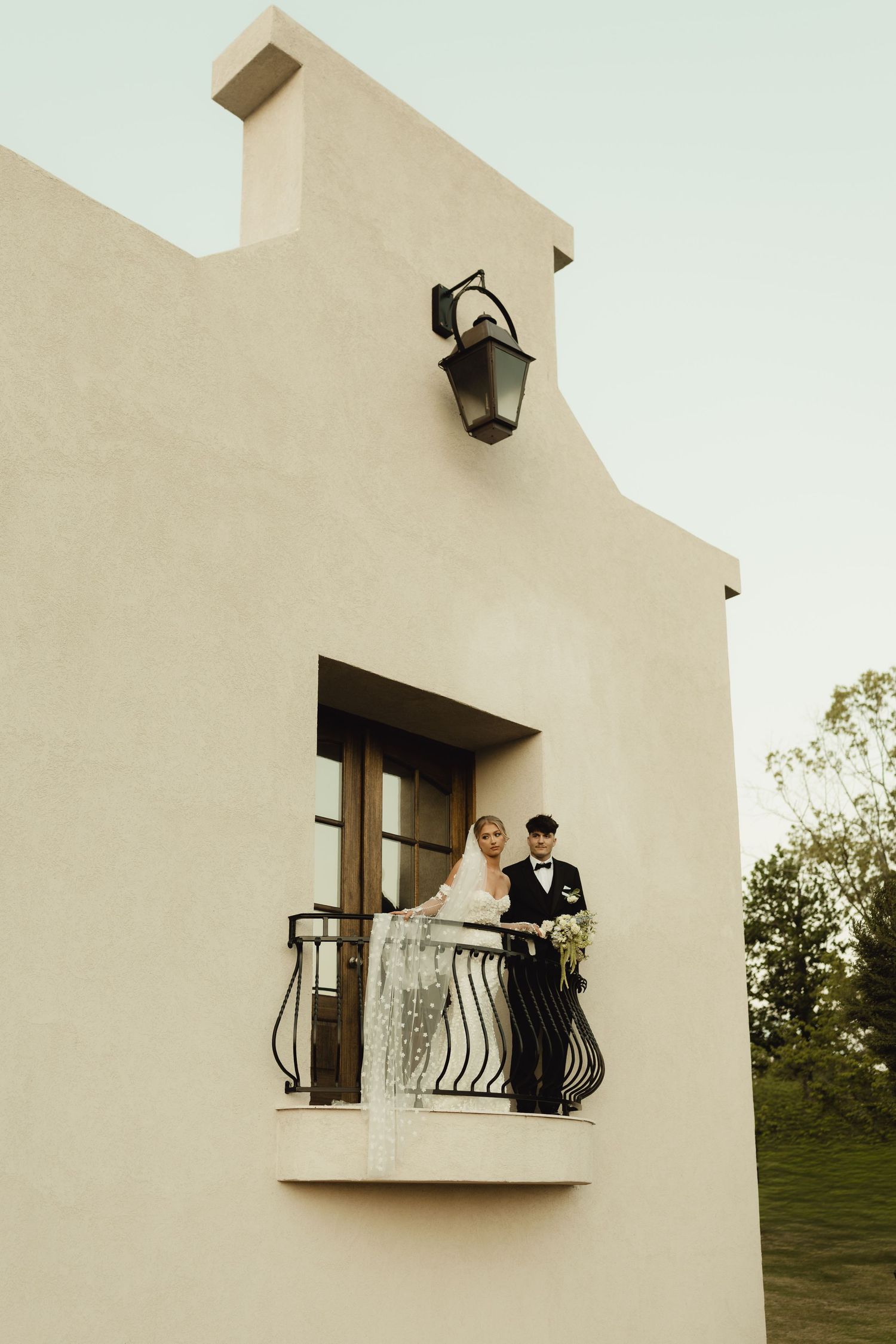 Bride and groom stand on a balcony of a beige stucco building, sharing a romantic wedding moment outdoors.