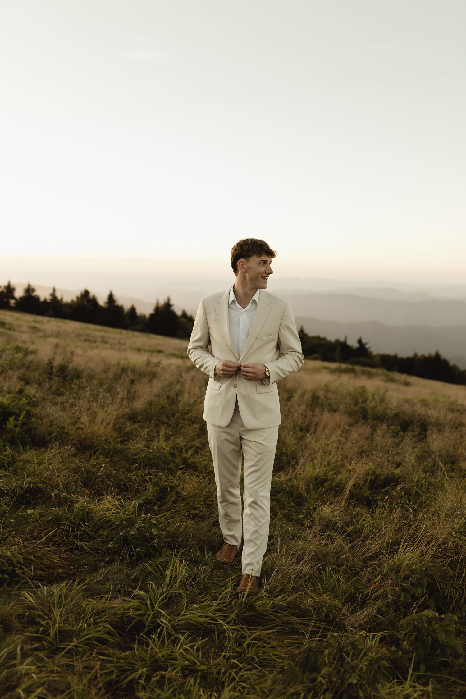 Young man in beige suit standing on a grassy mountain hillside at dusk with scenic mountain views.