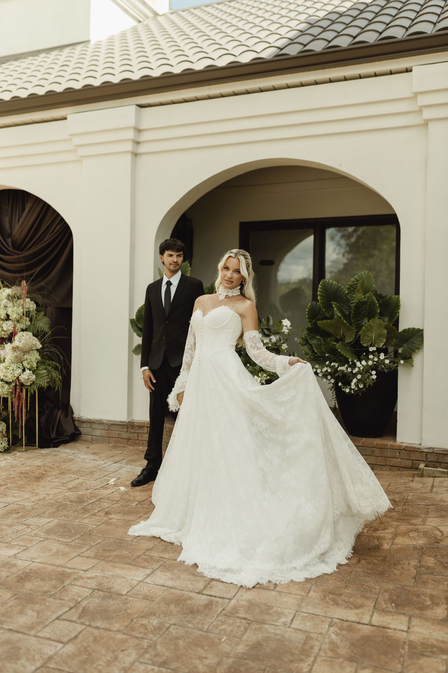 Bride in white lace off-shoulder gown twirls outside elegant venue as groom in black suit stands behind her.