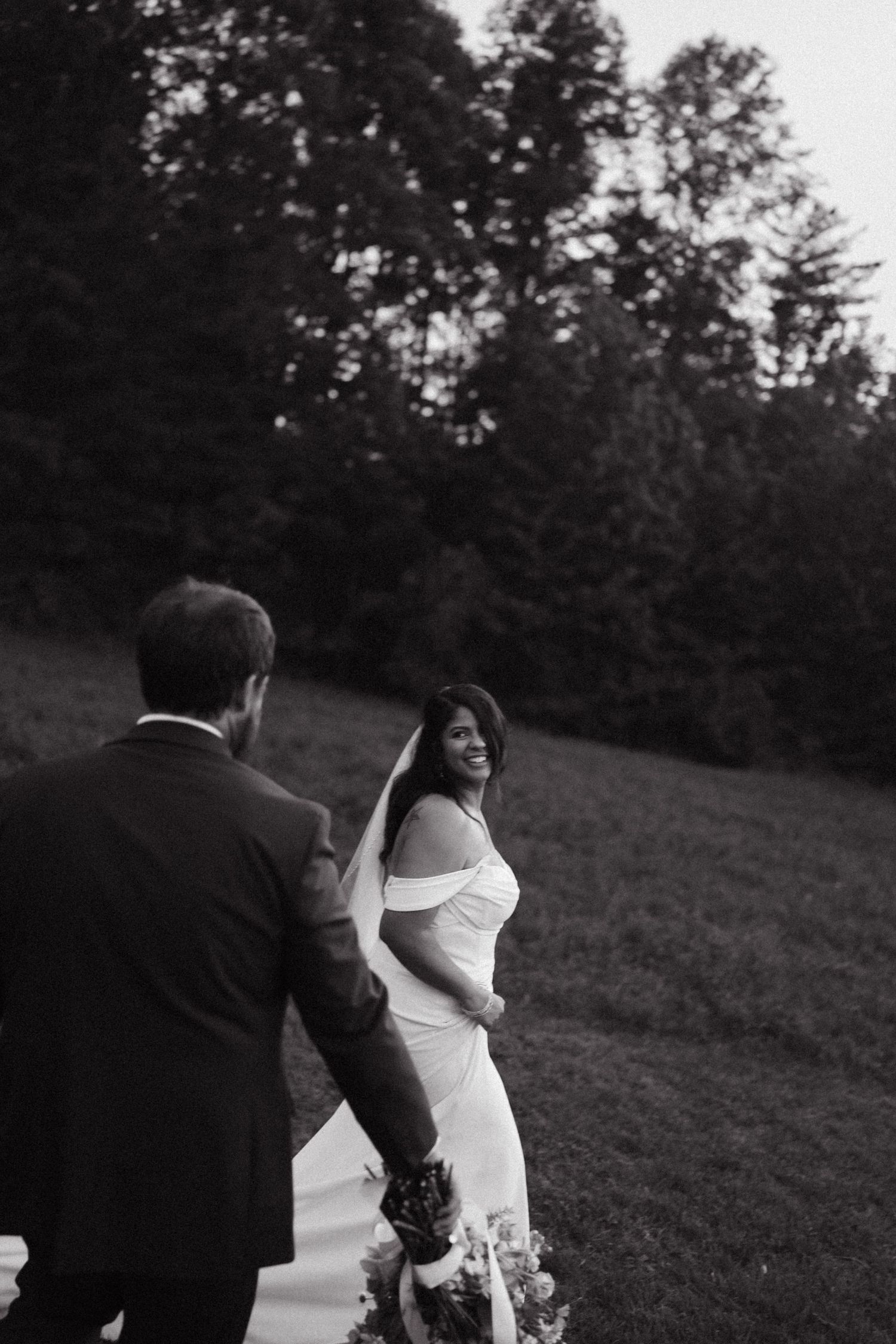 Black and white photo of a smiling bride in an off-shoulder gown holding hands with groom outdoors.
