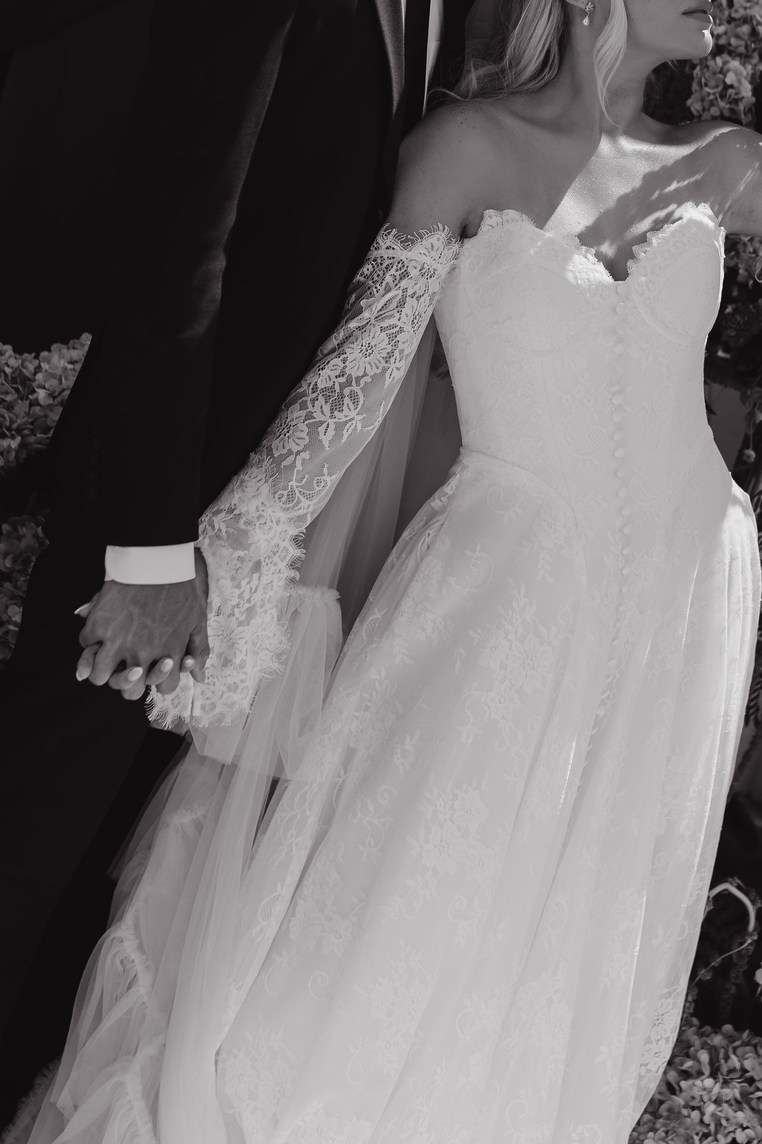 Black and white photo of bride in lace off-shoulder wedding gown holding hands with groom in dark suit.