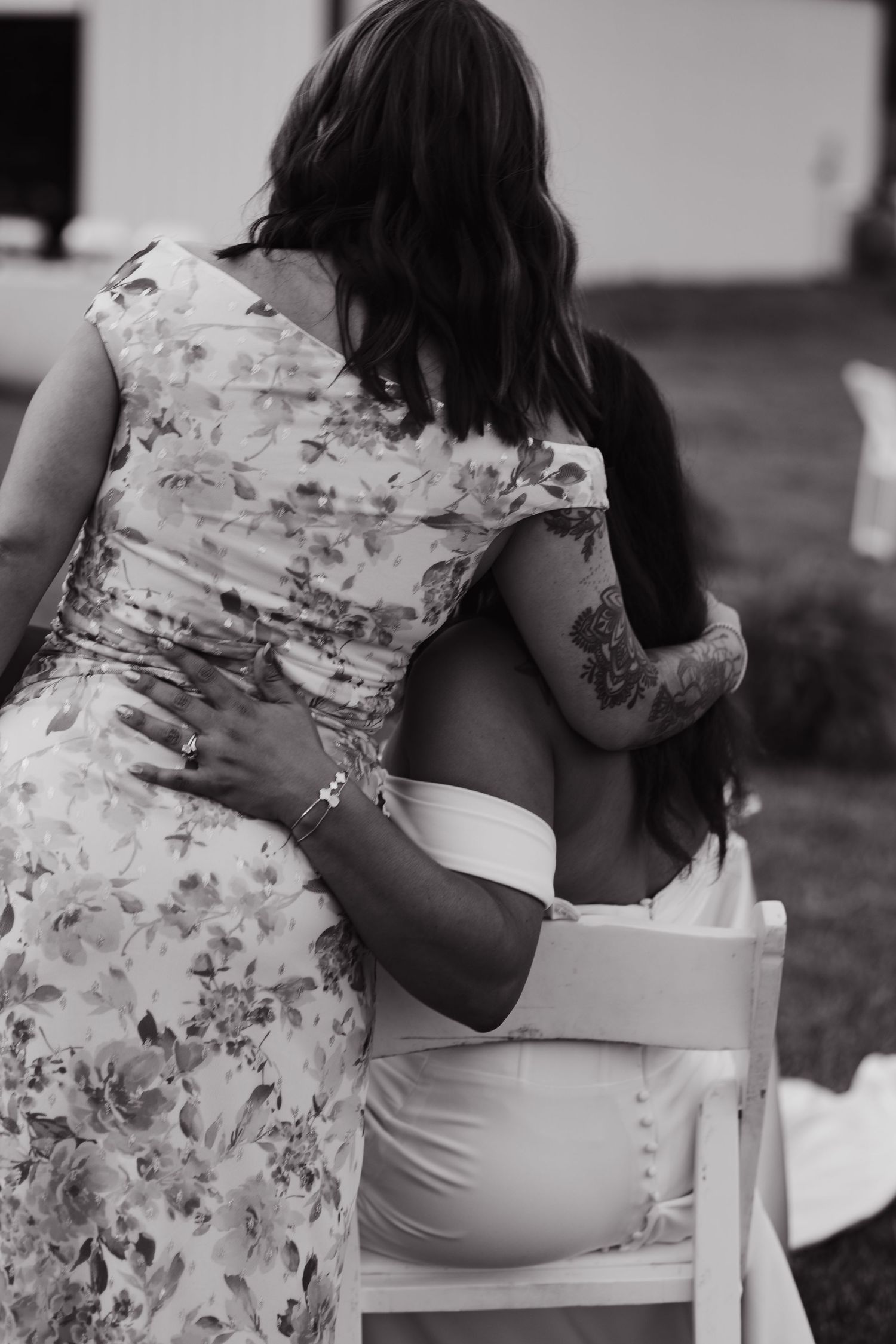 Black and white photo of two women embracing at an outdoor wedding, one seated in white dress with tattoos.