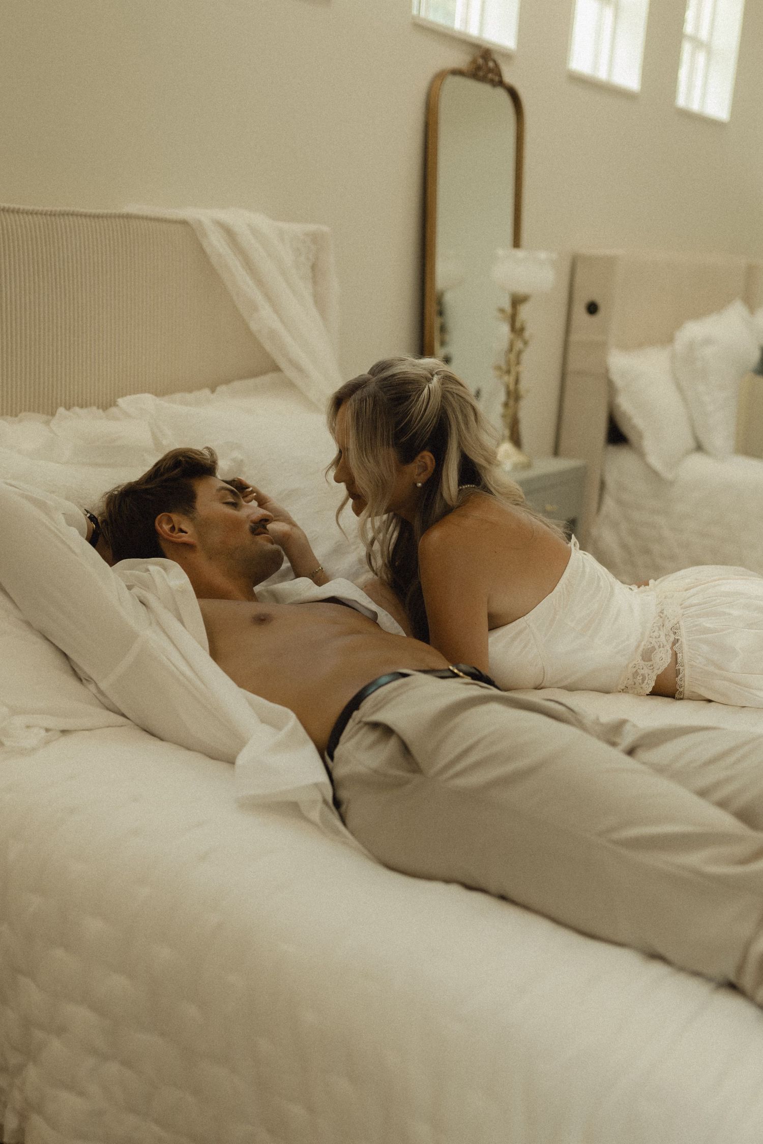 Couple in intimate bedroom setting, man shirtless lying on white bed while woman in white lingerie leans over him smiling.