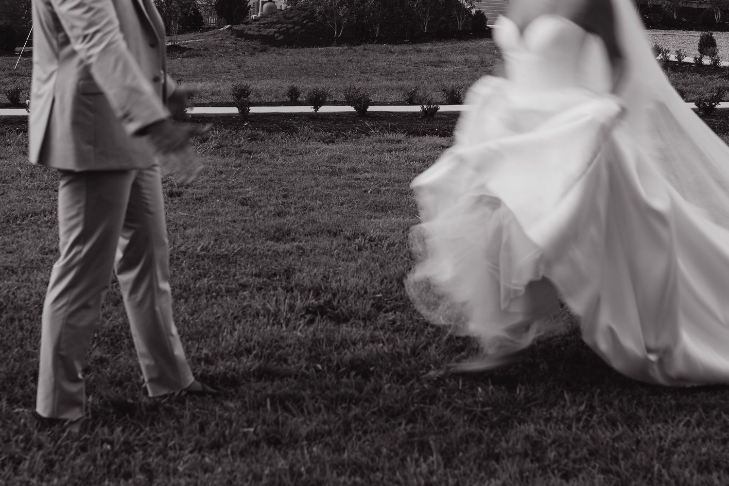 Black and white photo of a bride in flowing white gown and groom in suit walking on grass at outdoor wedding.