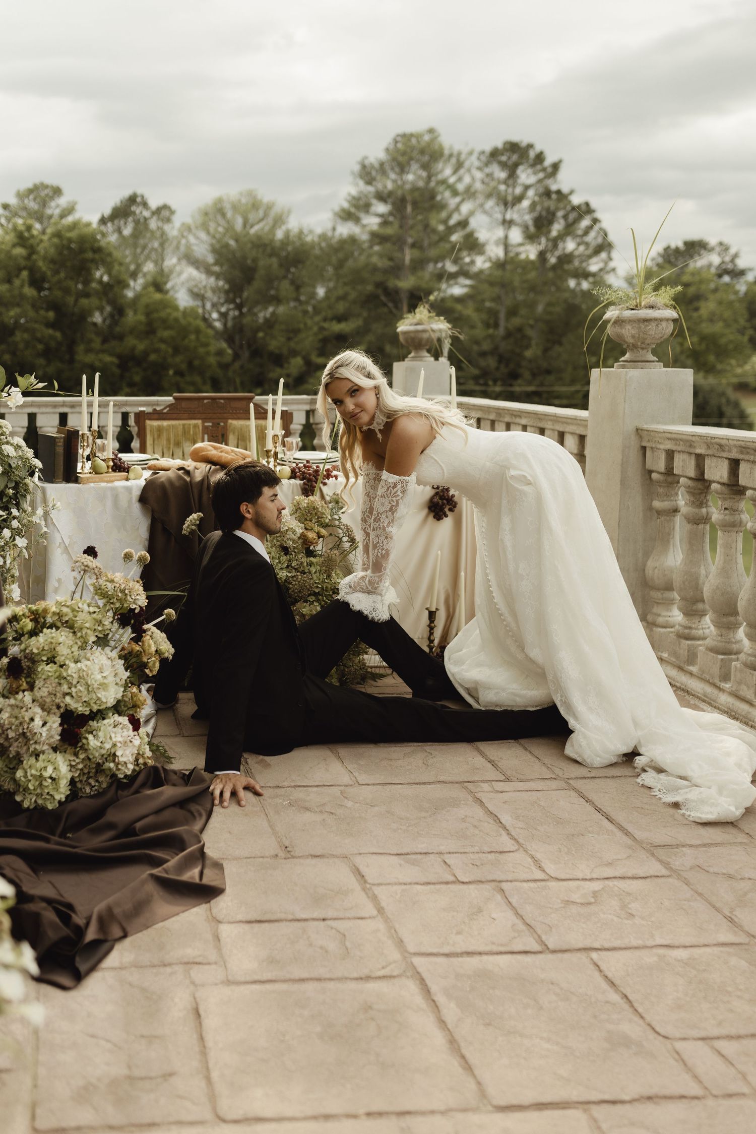 Bride in white gown sits on stone terrace beside groom in black suit, surrounded by lush greenery and floral arrangements.