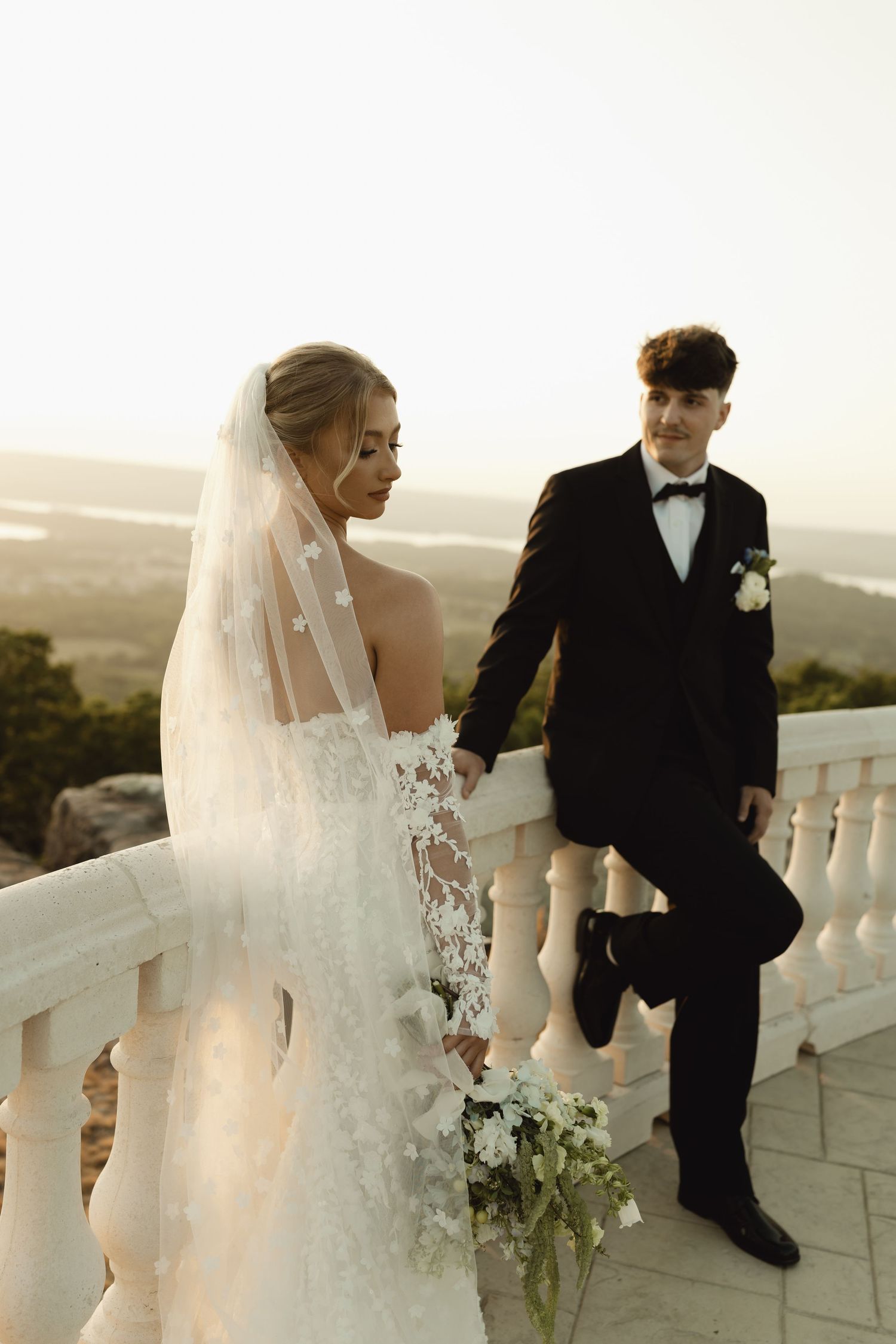 Bride in lace gown and veil holds groom's hand on scenic balcony at golden hour wedding.