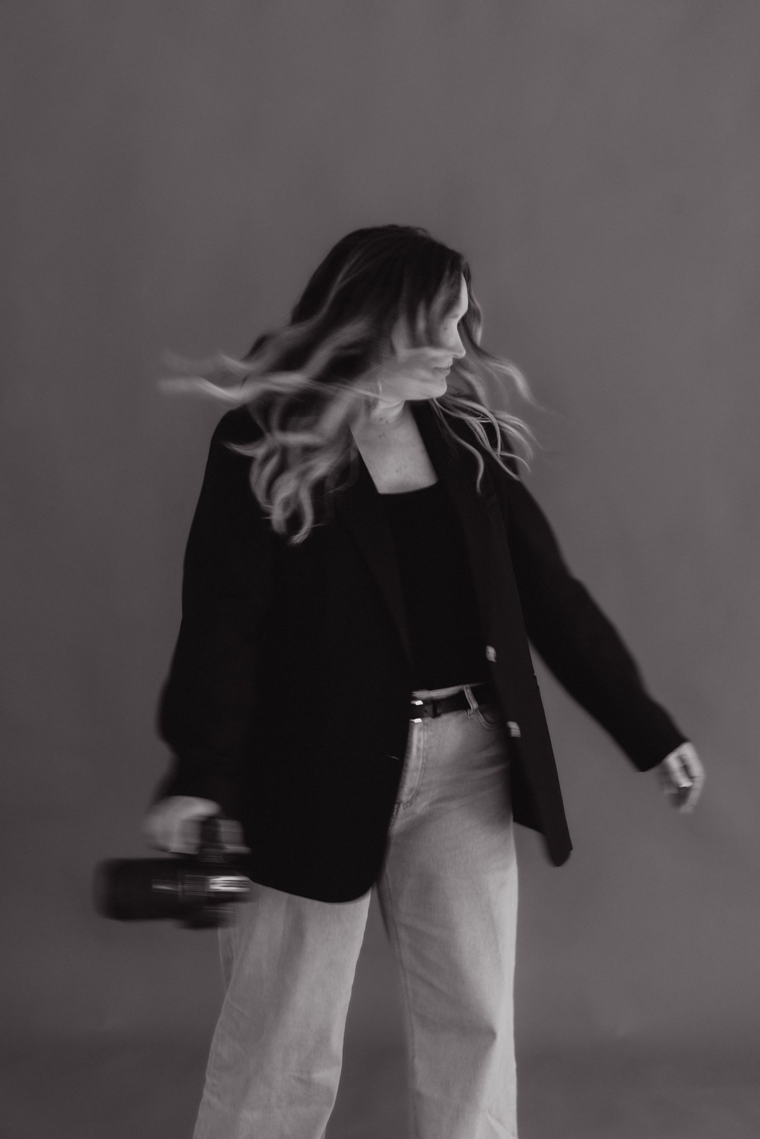 Black and white photo of a woman with flowing hair wearing a blazer and jeans, mid-motion in a studio setting.