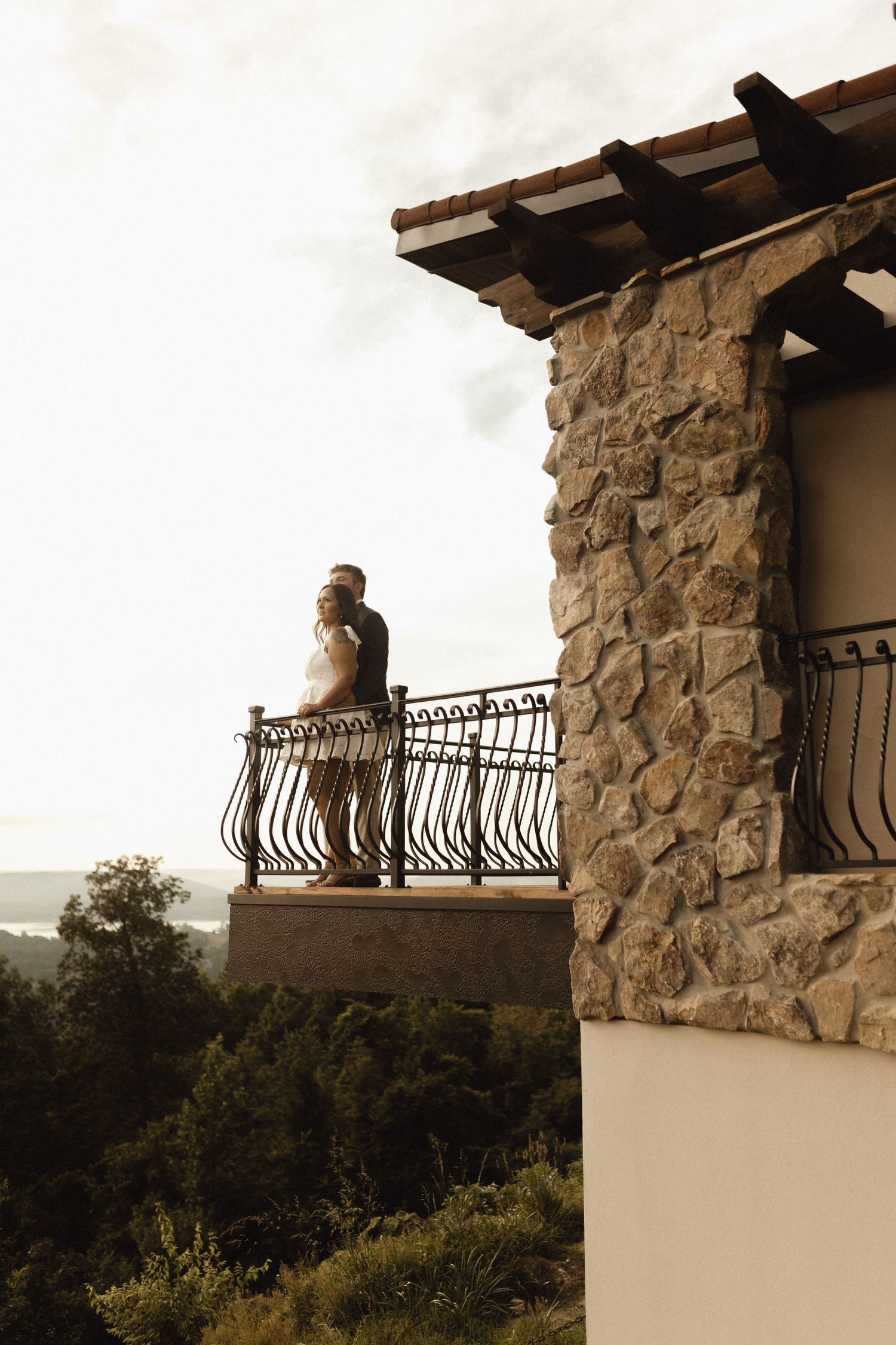 A figure stands on a rustic stone balcony overlooking a scenic mountain landscape at sunset.