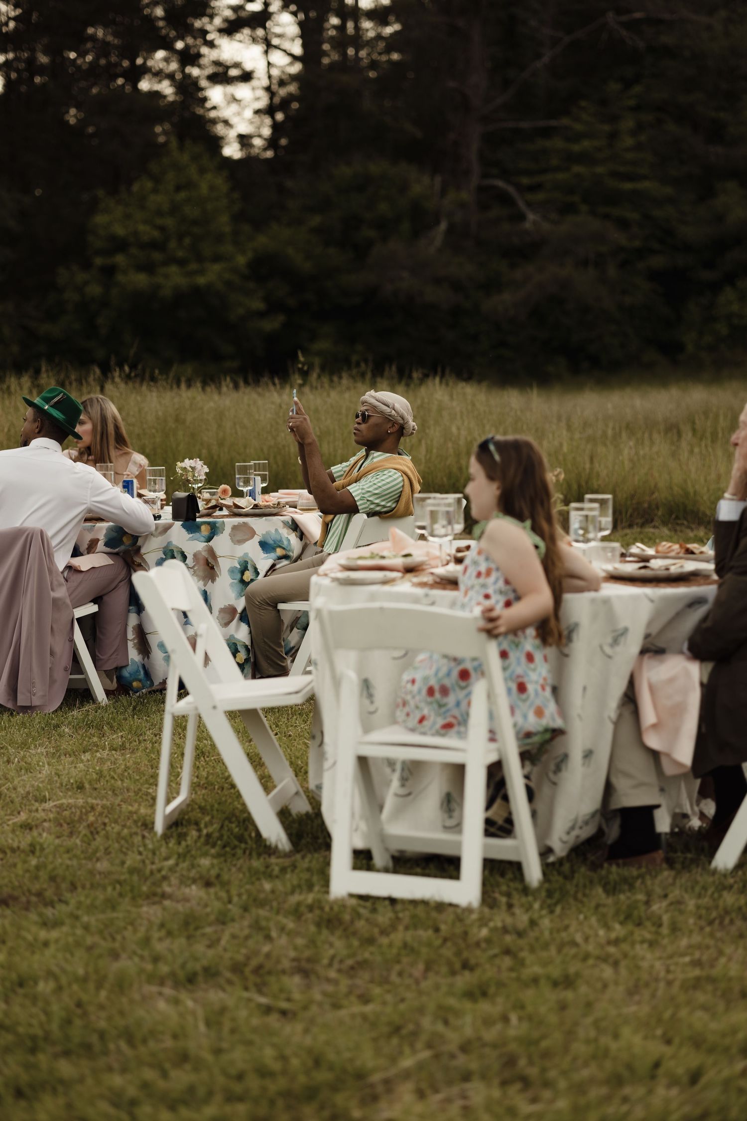 Diverse group of guests dining at an elegant outdoor garden party table surrounded by tall grass and trees.