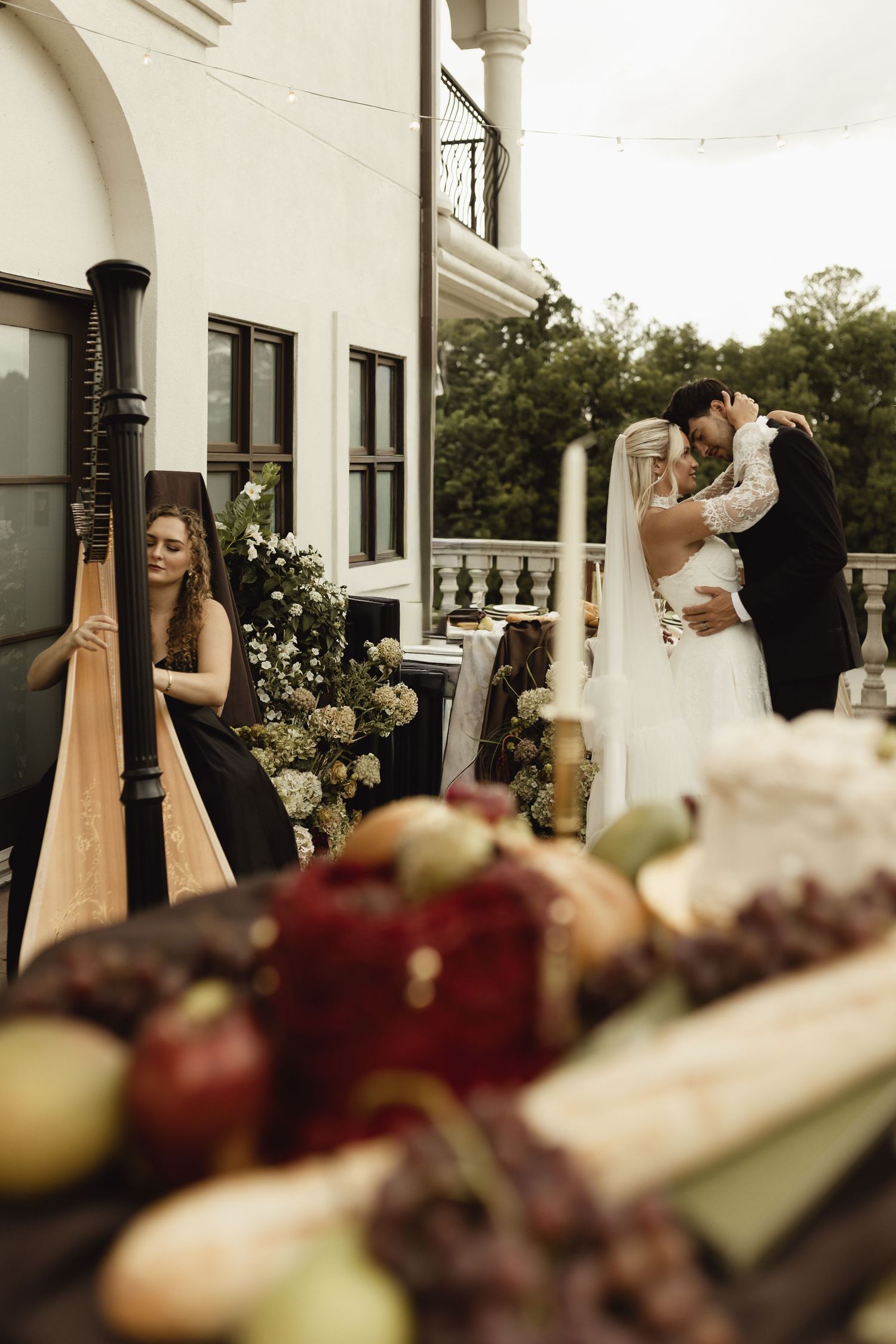 Bride and groom share a romantic moment at outdoor wedding reception with harpist and elegant grazing table in foreground.