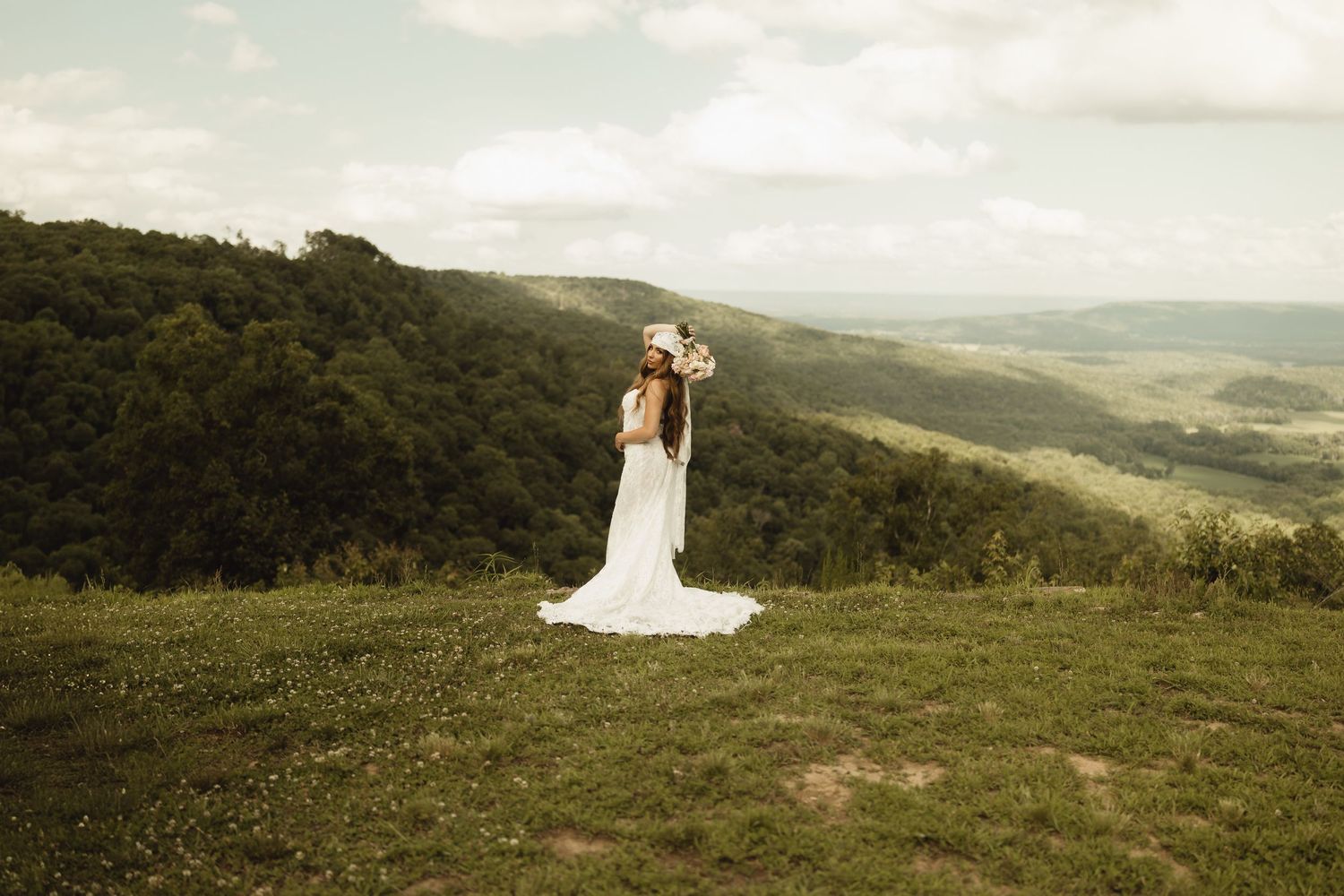 Bride in white wedding dress with floral crown standing on green hilltop overlooking scenic valley landscape.