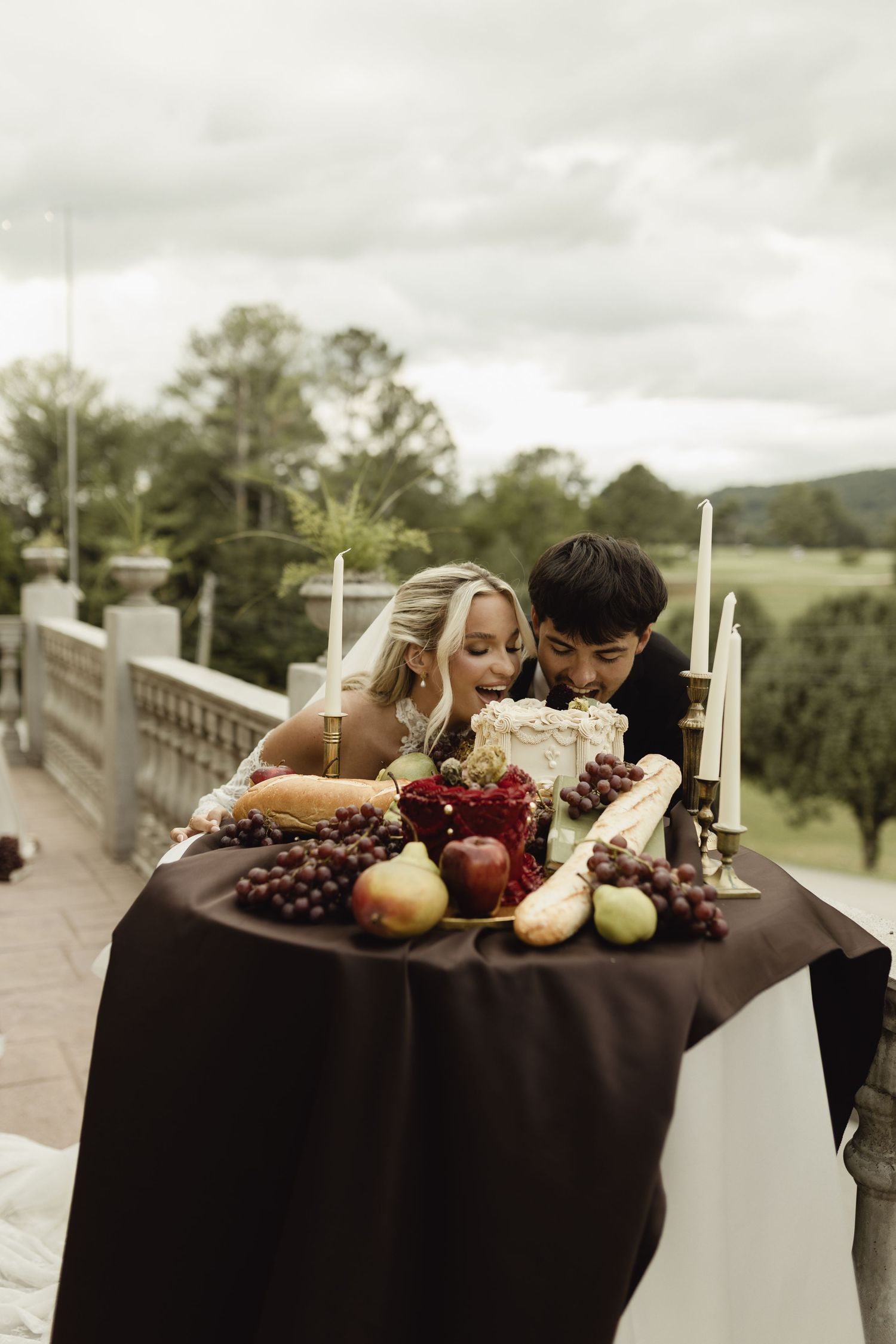 Bride and groom leaning over elegant outdoor wedding table with fruit, candles, and white cake on cloudy day.