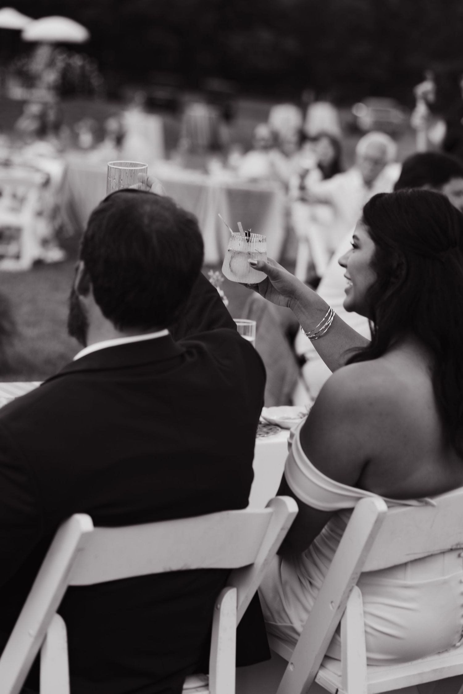 Black and white photo of a couple at an outdoor wedding reception, woman in strapless dress raising a cocktail glass.