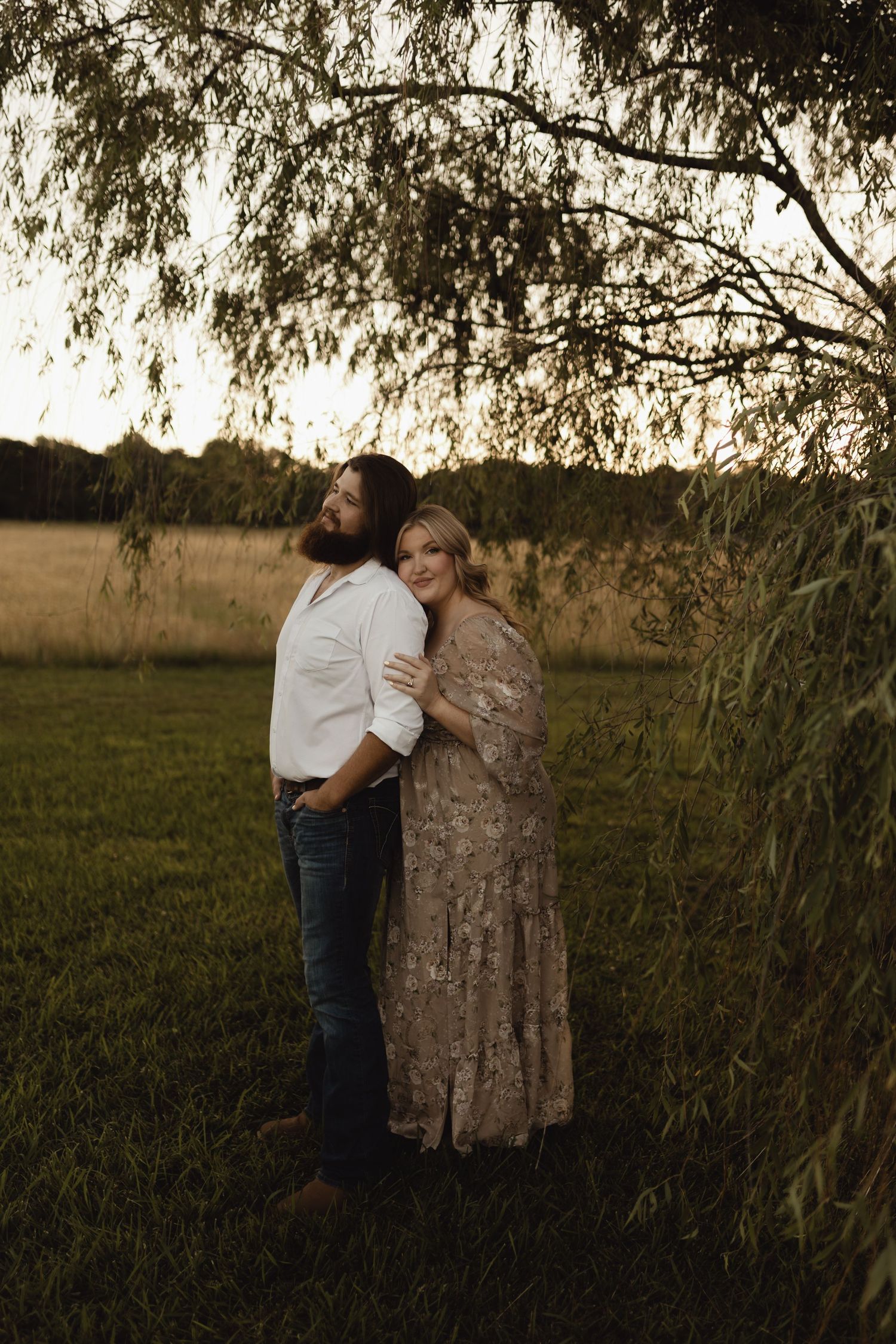 A couple embraces under willow tree branches in a golden field at sunset, woman in floral dress smiling warmly.