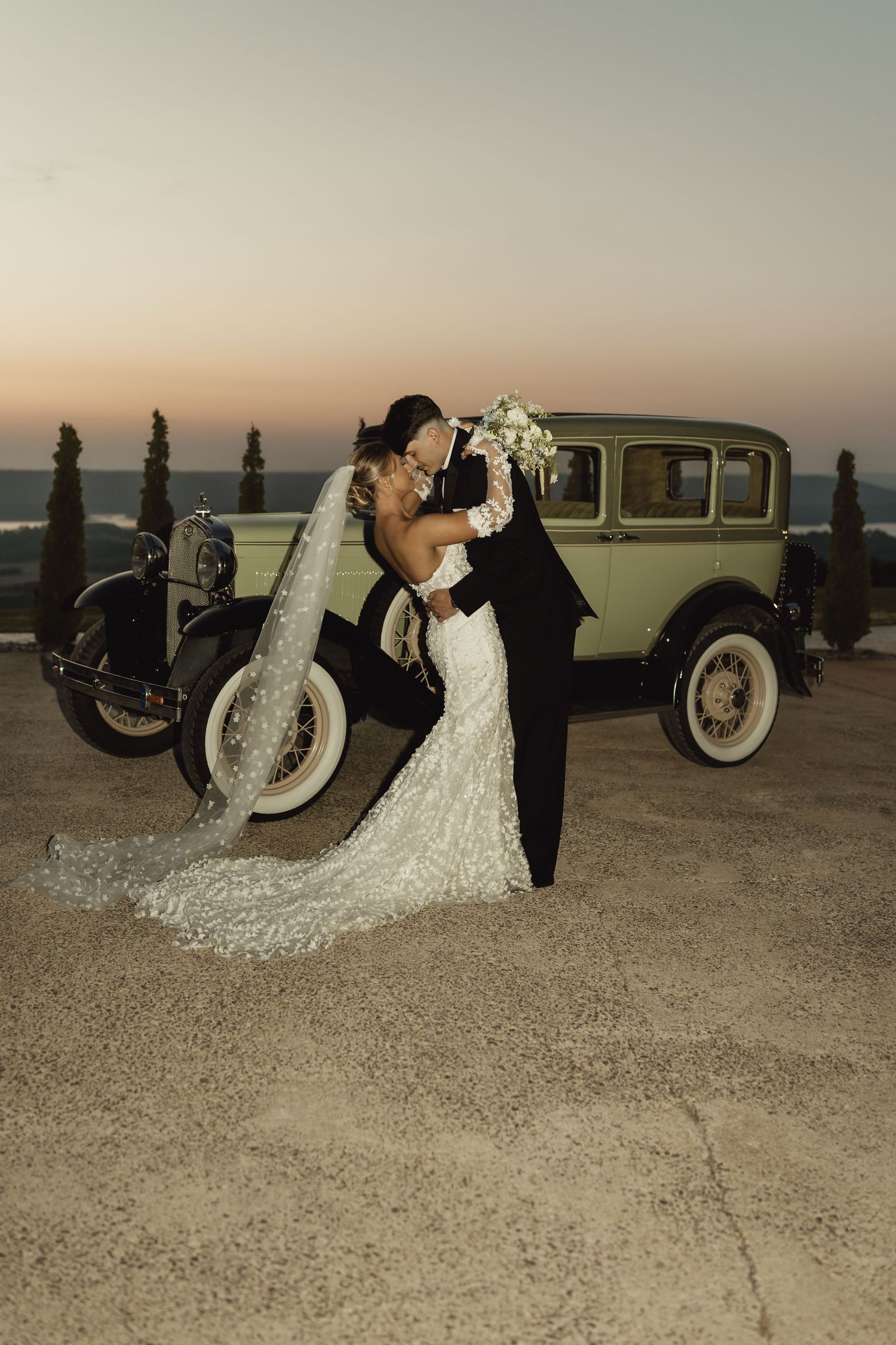 Bride and groom kissing beside a vintage classic car at sunset, bride in lace gown with long veil holding white bouquet.