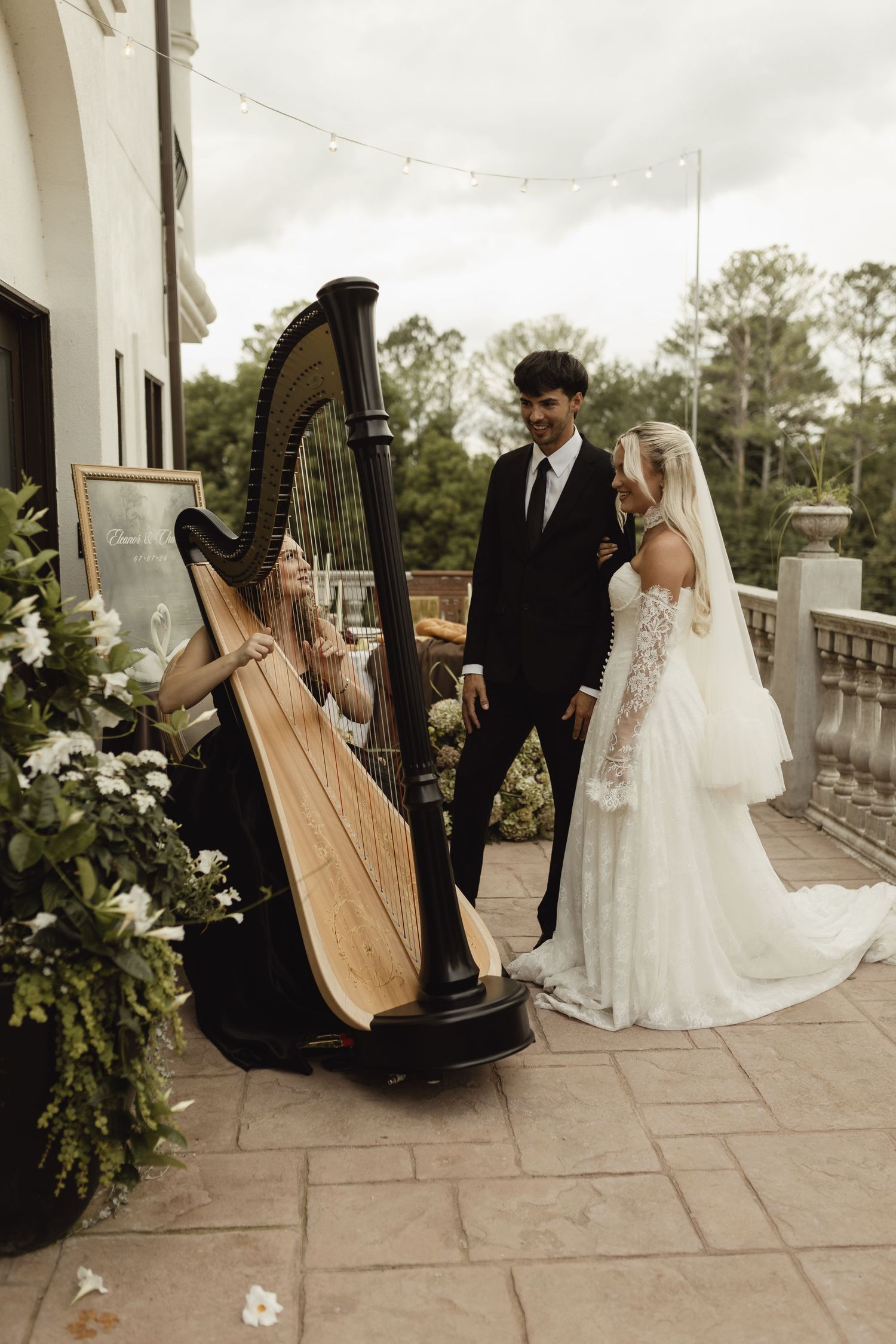 Bride and groom listen to a harpist playing at their outdoor wedding reception on an elegant terrace.