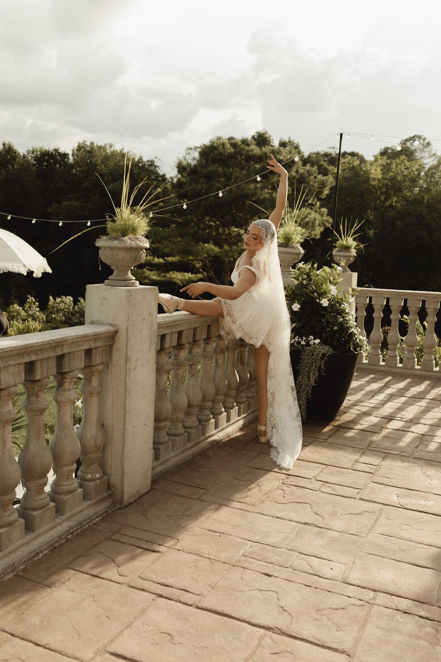 Bride in lace wedding dress and veil performs elegant ballet pose on stone terrace surrounded by lush greenery.