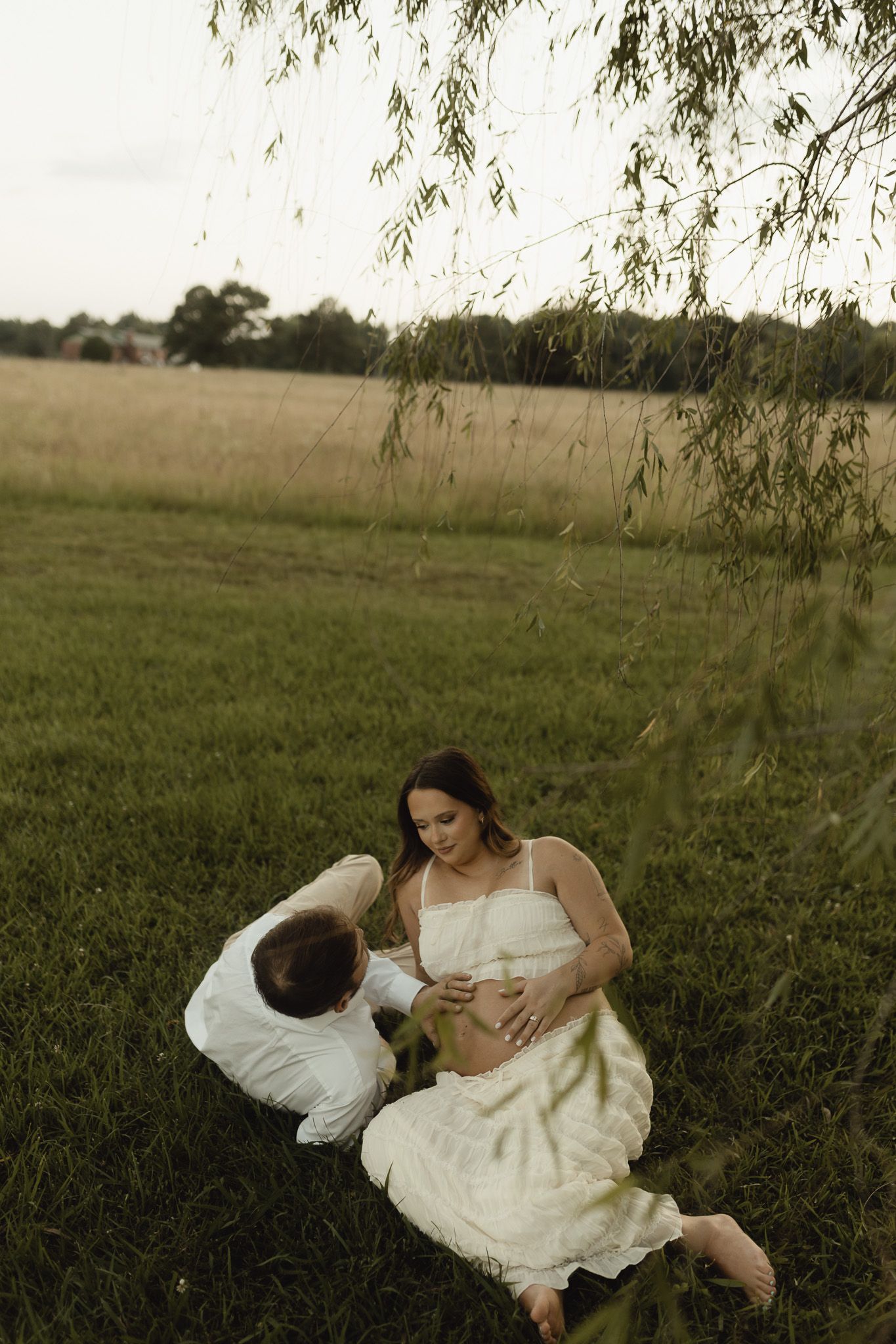 Pregnant couple sitting under willow tree in green field, woman in white dress cradling baby bump at golden hour.