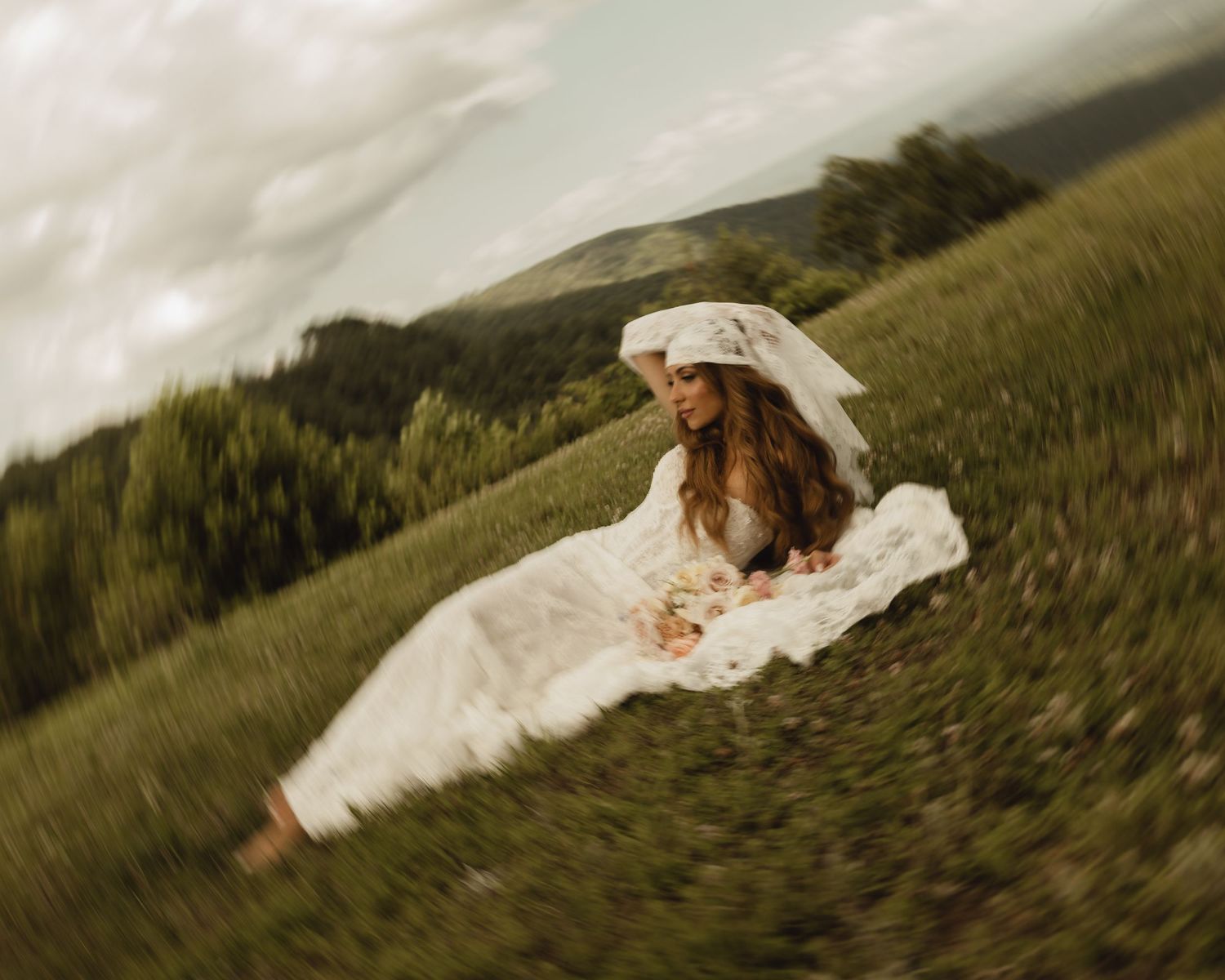 A bride in a white gown and veil sits gracefully in a lush green meadow with misty mountains in the background.