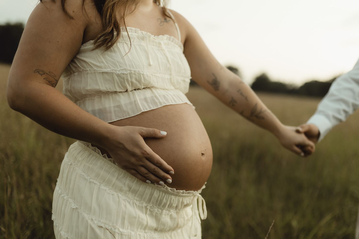 Pregnant woman in white dress holding baby bump in a field, holding hands with partner at golden hour.