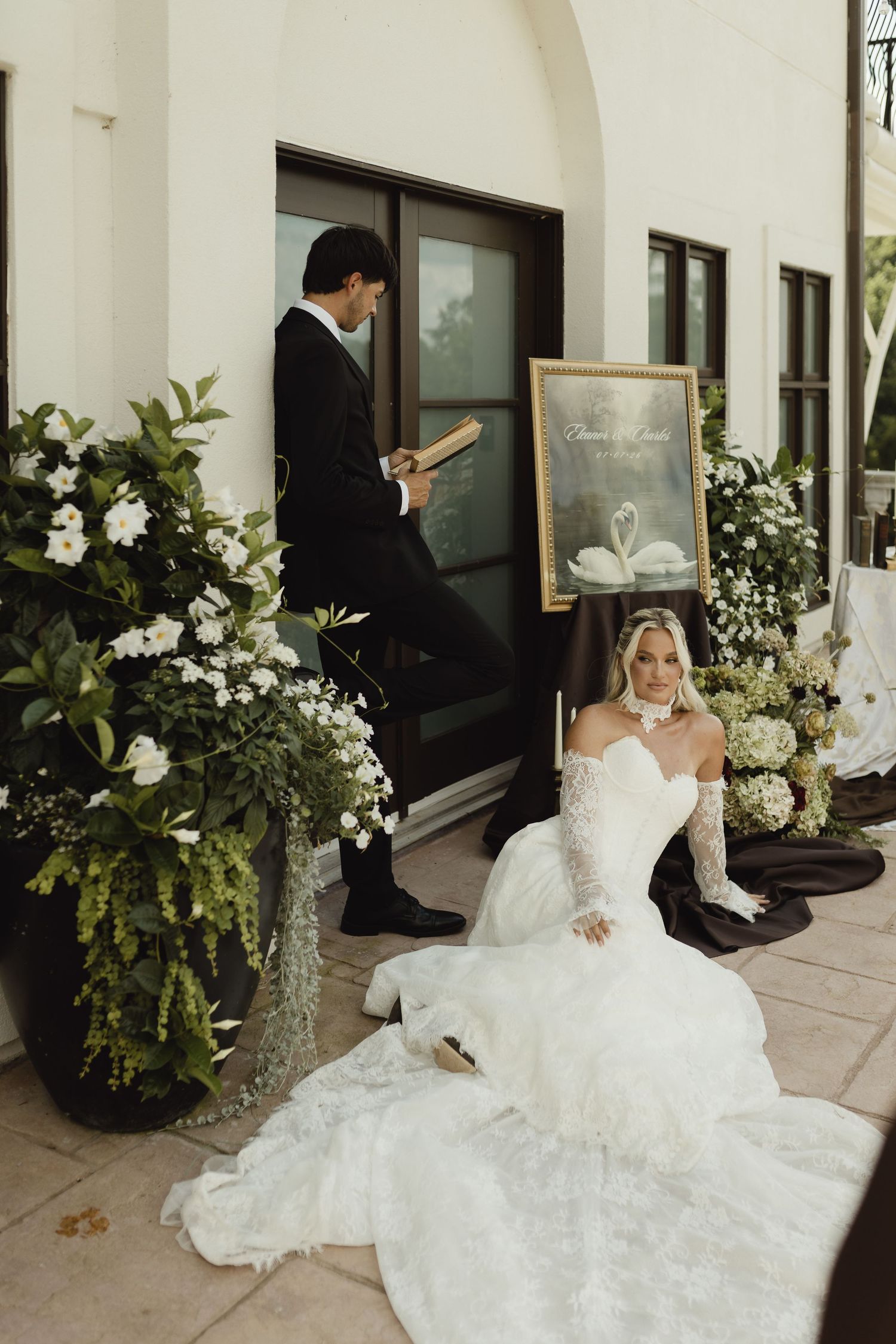 Bride in white off-shoulder gown sits on steps beside lush floral arrangements as groom reads nearby.