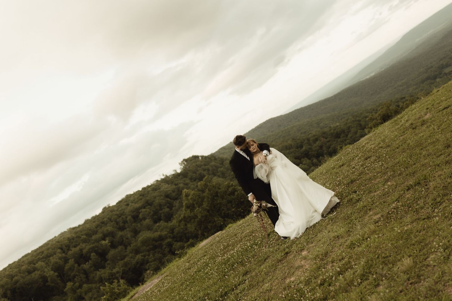 A bride and groom share a romantic kiss on a lush green hillside with scenic mountain views on their wedding day.