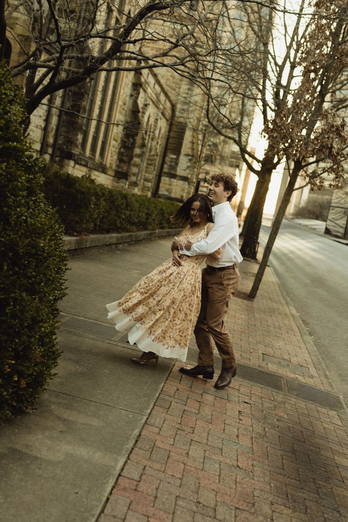 Couple dancing joyfully on a brick sidewalk, woman in floral dress, man in white shirt and brown pants, urban setting.