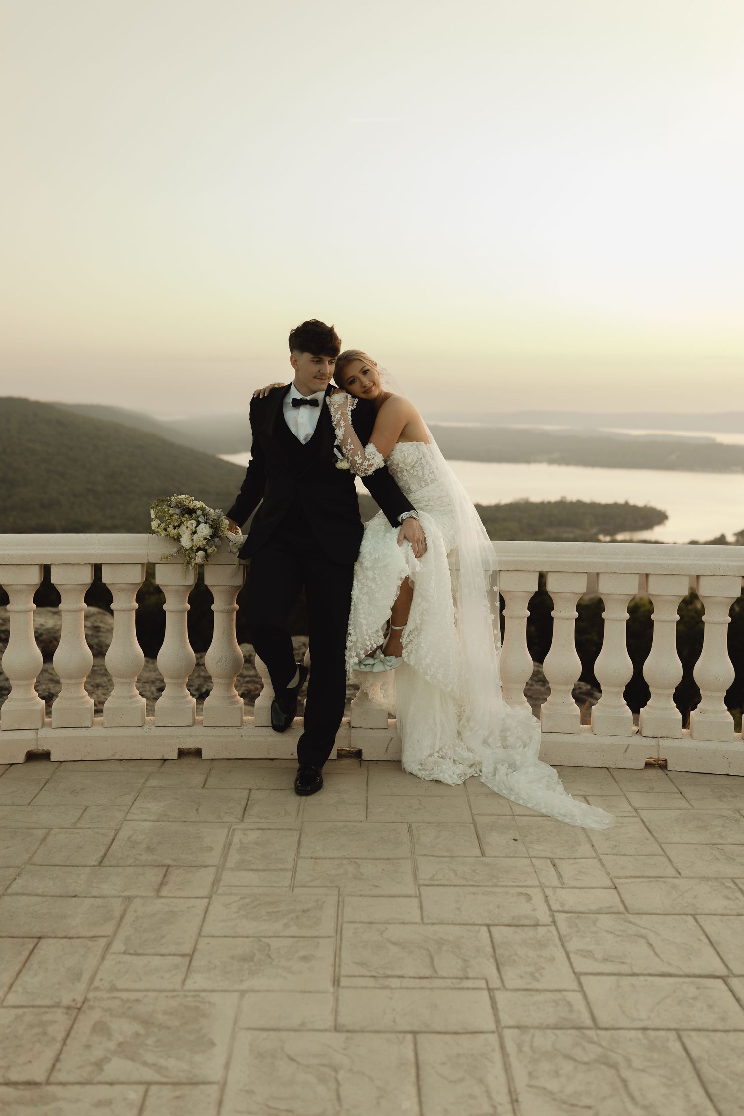 Bride and groom pose on a scenic terrace overlooking a lake at sunset, she in lace gown, he in black tuxedo.