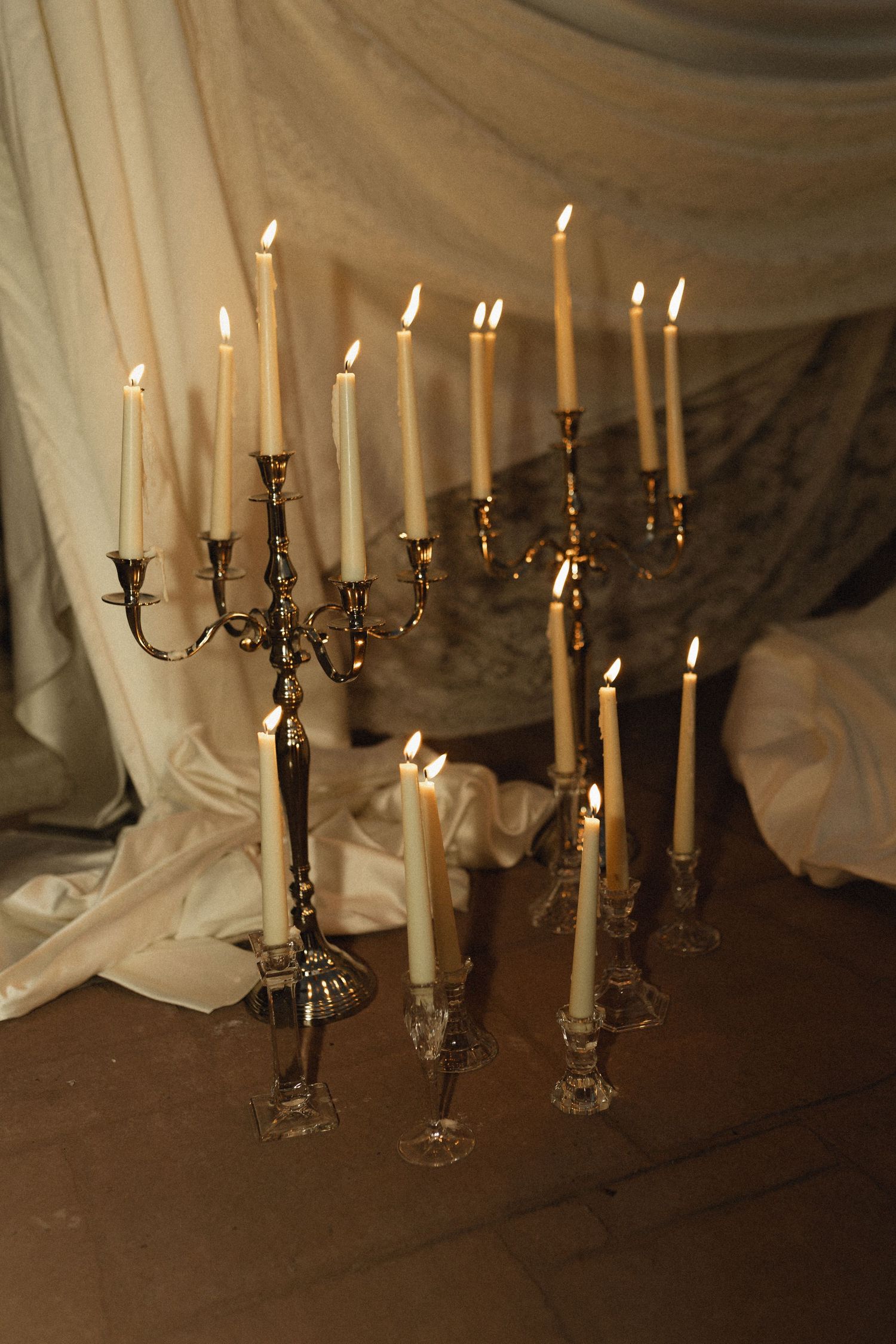 Elegant silver candelabras with lit white taper candles arranged on a wooden floor against draped white fabric backdrop.
