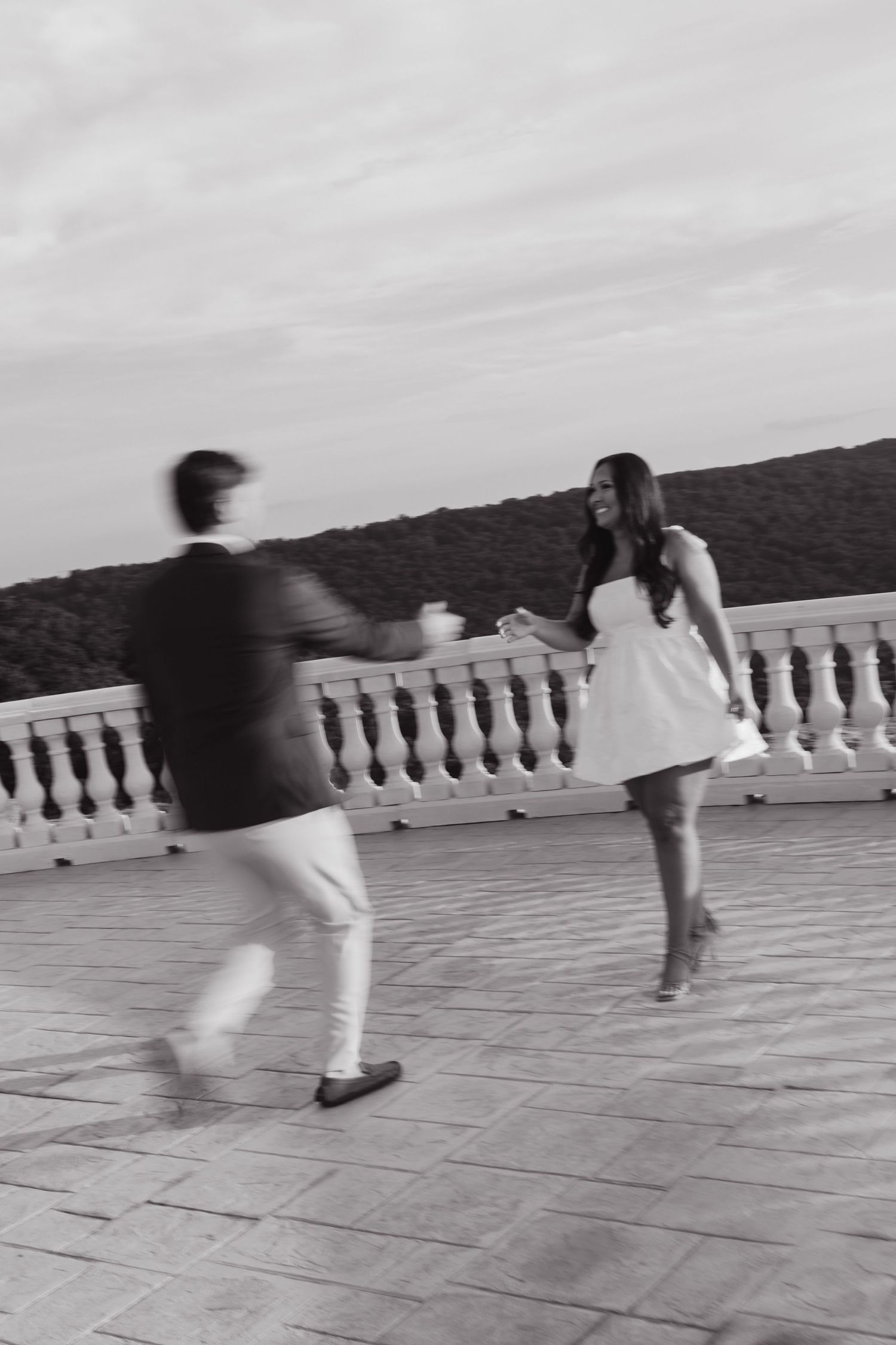 A couple dancing together on a terrace with white balustrades against a cloudy sky in black and white.