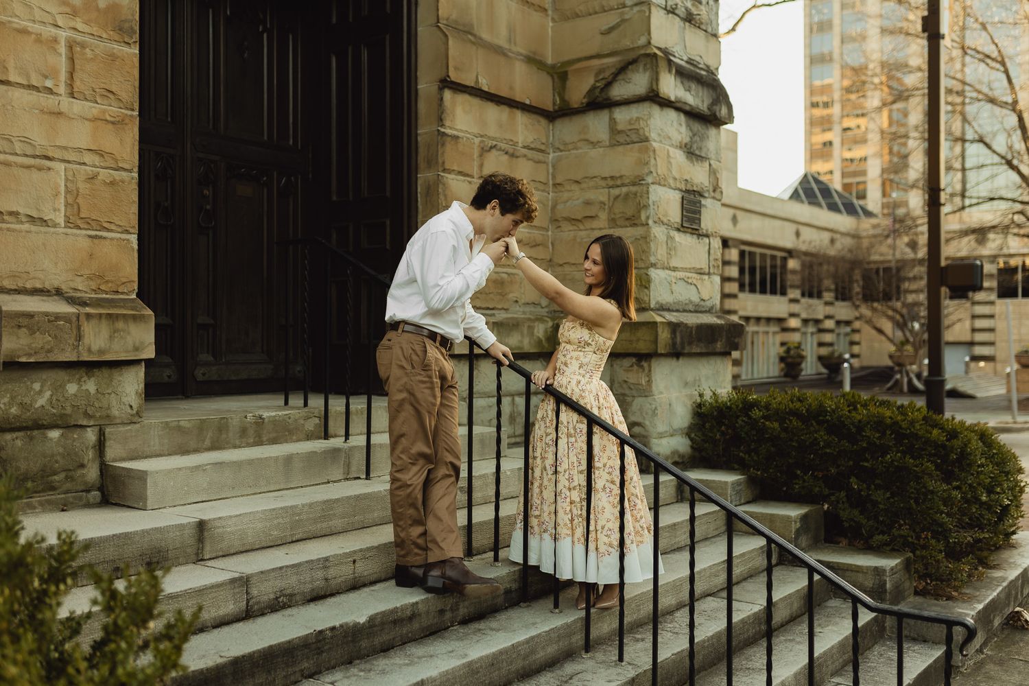 Couple sharing a romantic moment on stone church steps, woman in floral dress holding man's face, urban setting.
