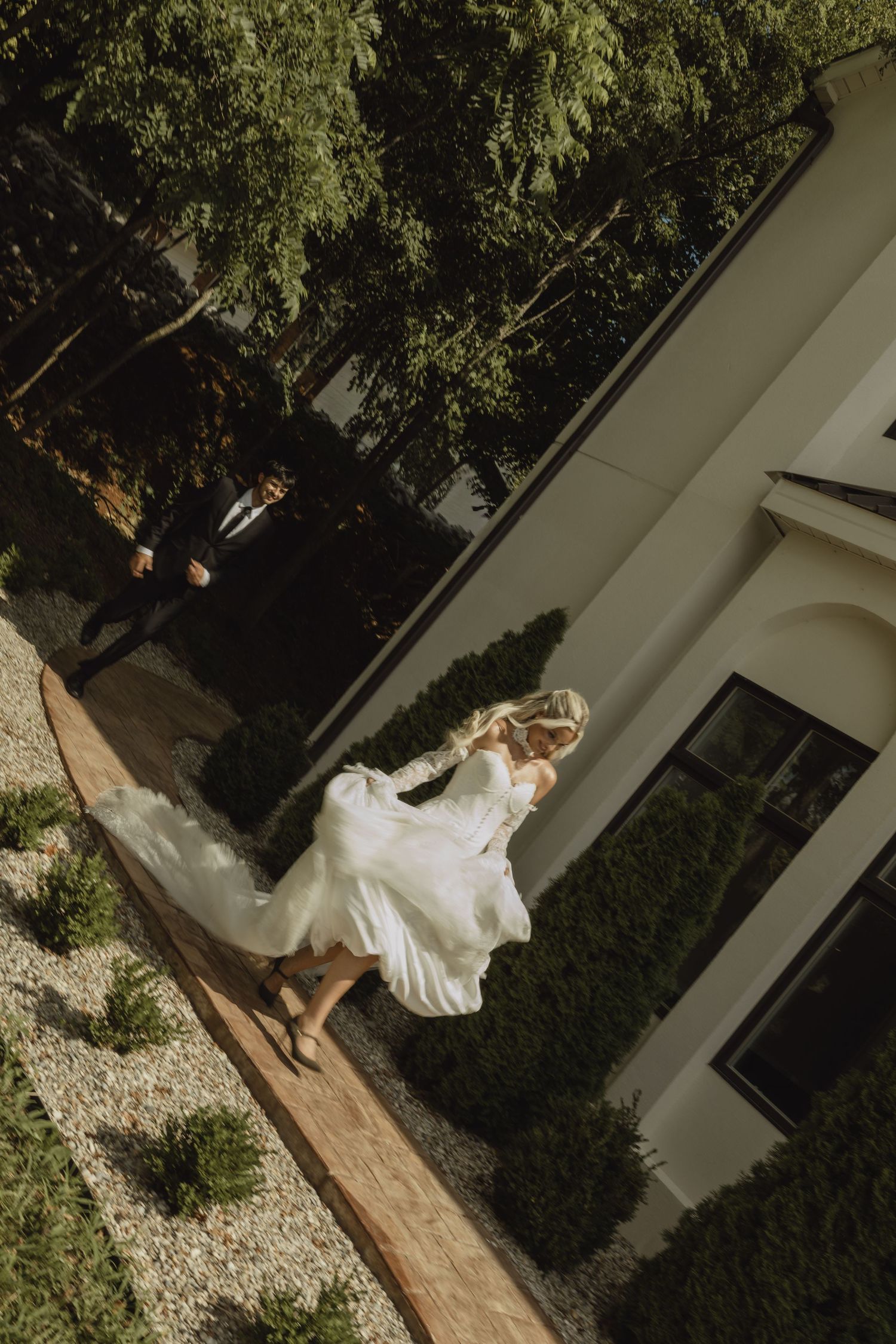 Aerial view of a bride in a flowing white wedding dress twirling outside a modern white building surrounded by greenery.