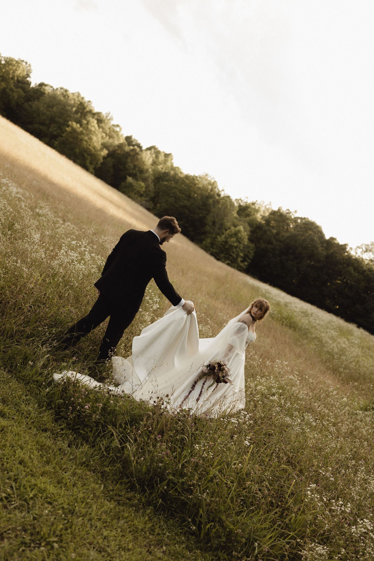 A groom in a black suit helps his bride in a flowing white gown walk up a lush green hillside meadow.