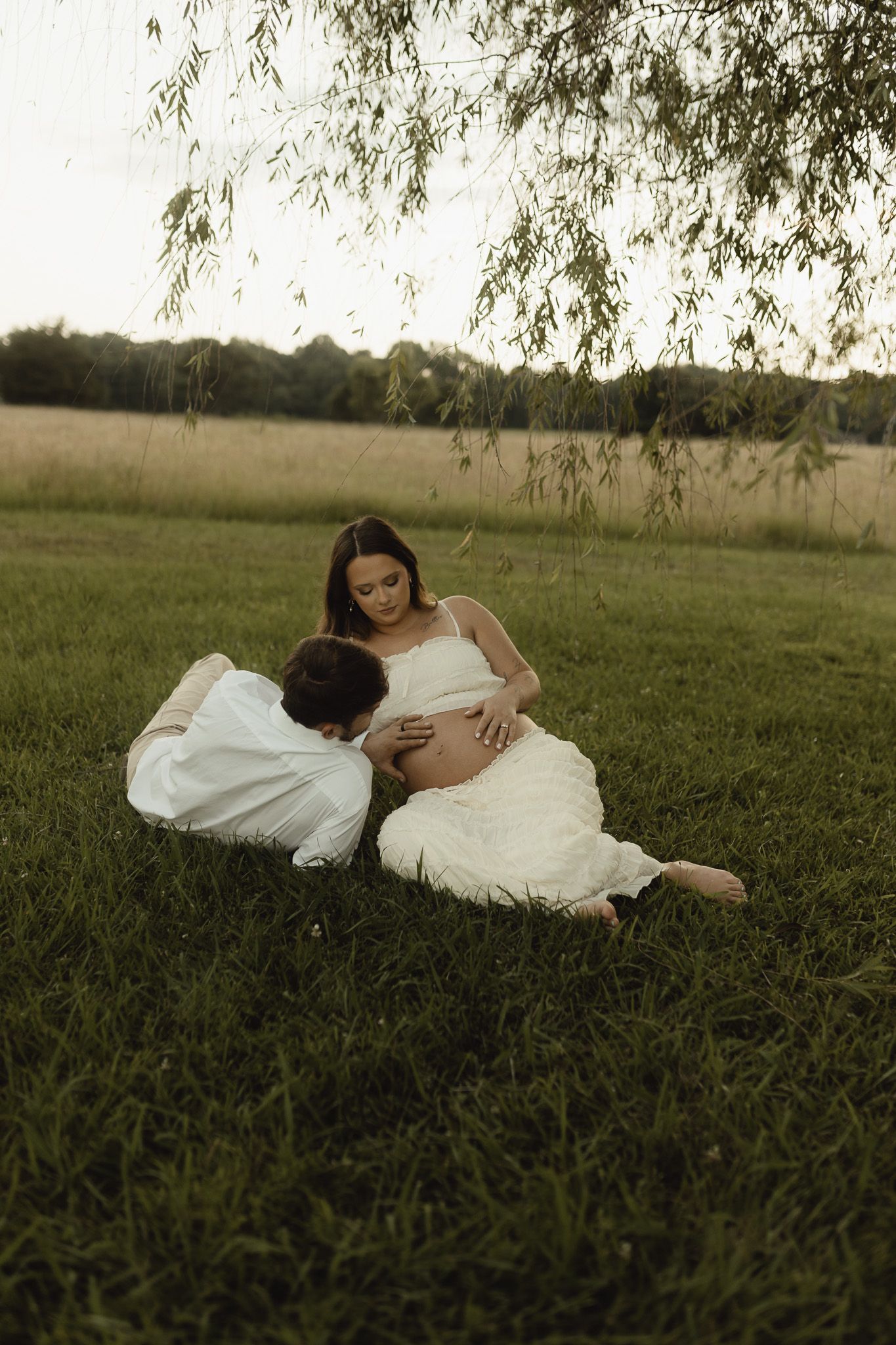 A couple sits on lush green grass under a willow tree, the man kissing a pregnant woman's belly in a serene outdoor setting.