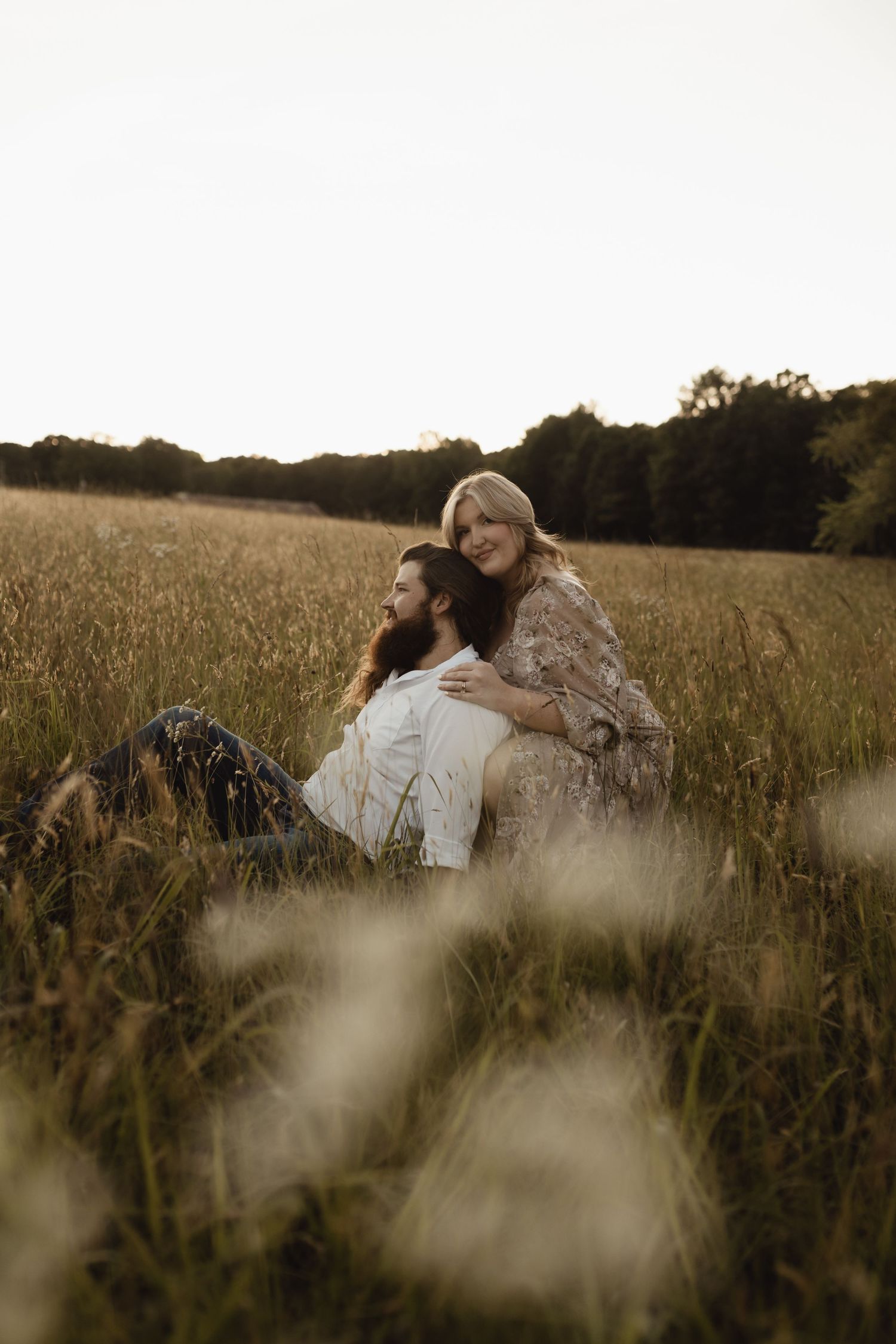 A couple sits together in a golden field, the woman embracing the man from behind as they enjoy a serene outdoor moment.