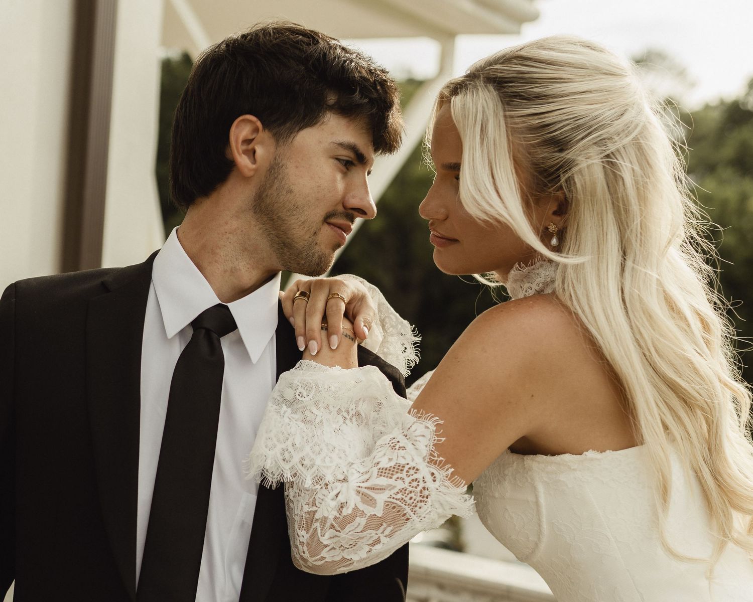 A bride and groom in wedding attire share an intimate moment, the bride in lace sleeves and the groom in a black suit.