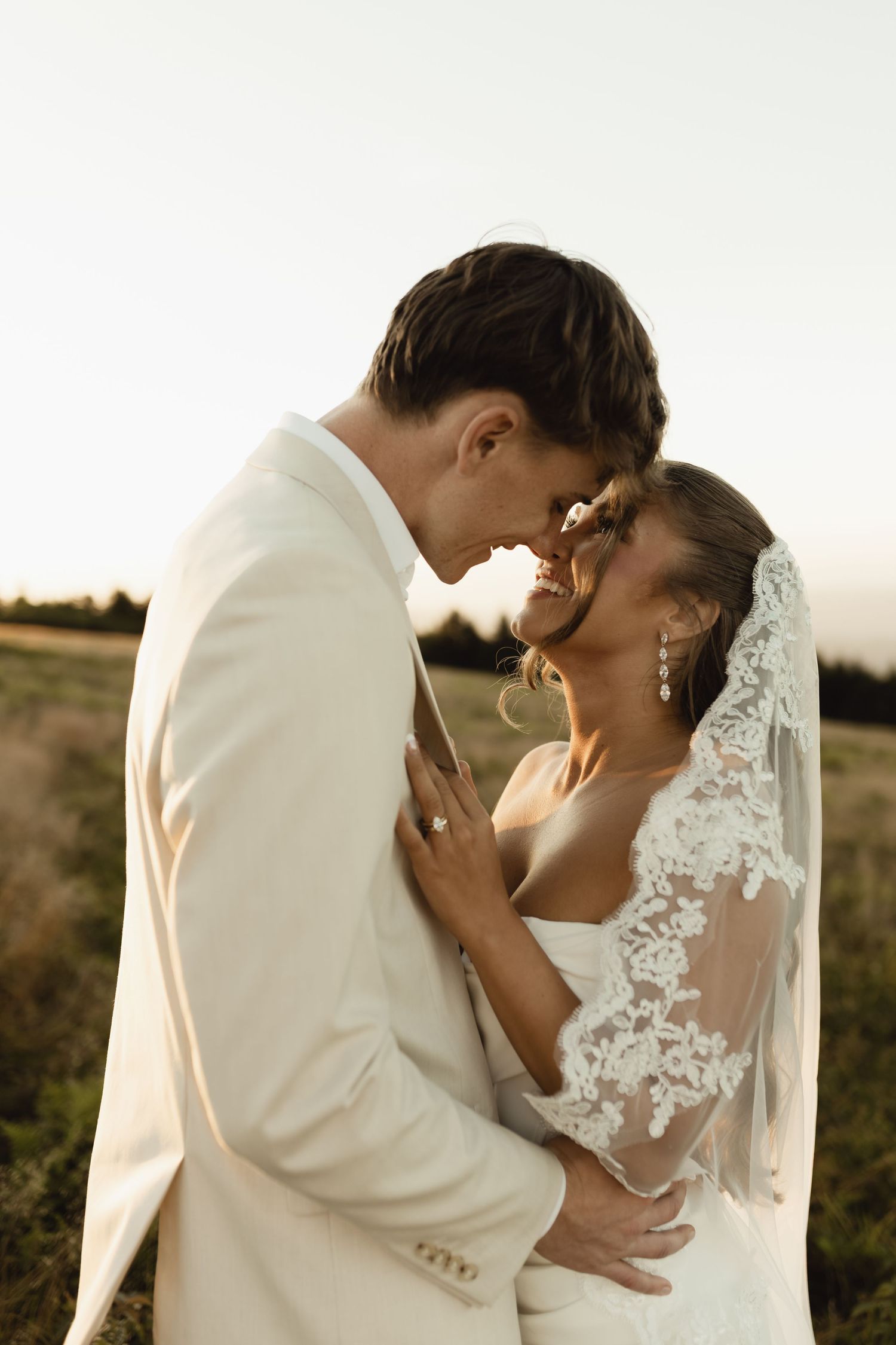 A bride and groom share a romantic moment in a golden field, with the bride wearing a lace veil and the groom in a white suit.