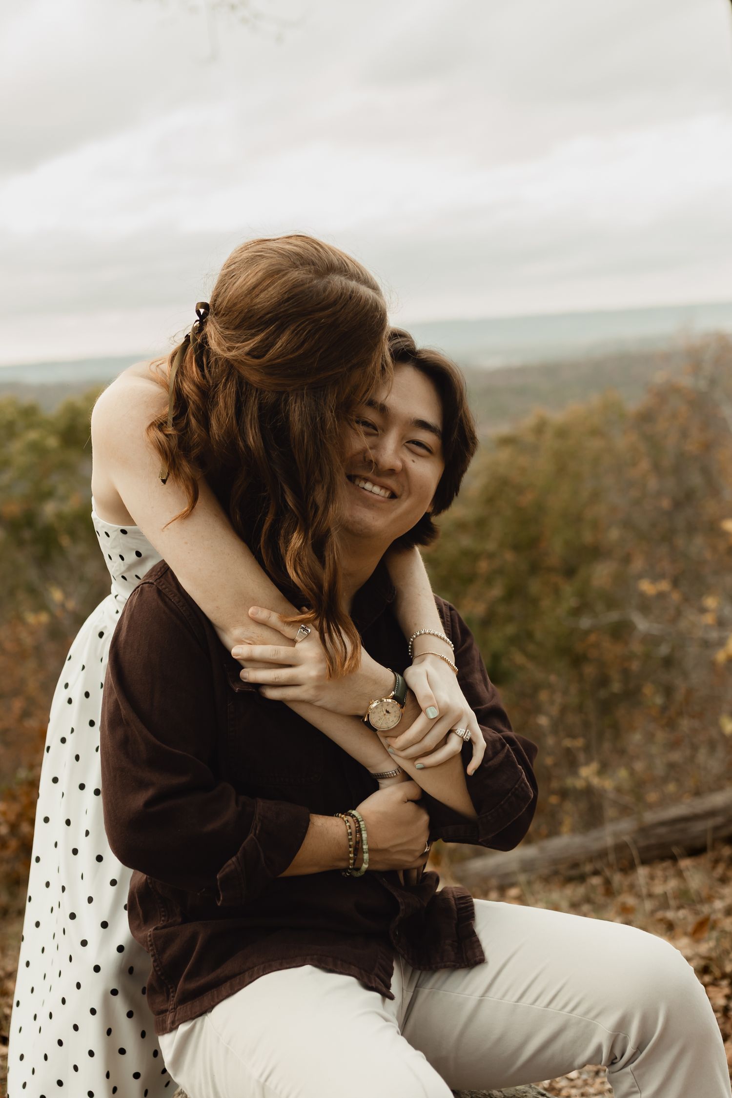 A couple embraces warmly outdoors during an autumn day with a natural landscape in the background.