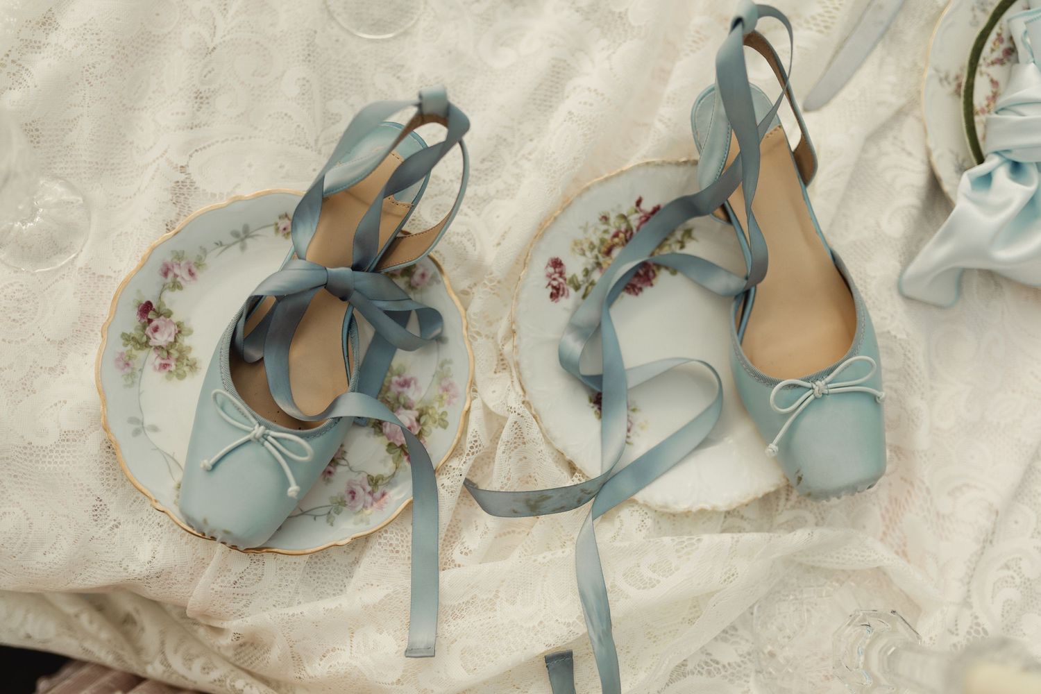 Elegant light blue ballet flats with satin ribbons displayed on floral china plates and lace fabric.