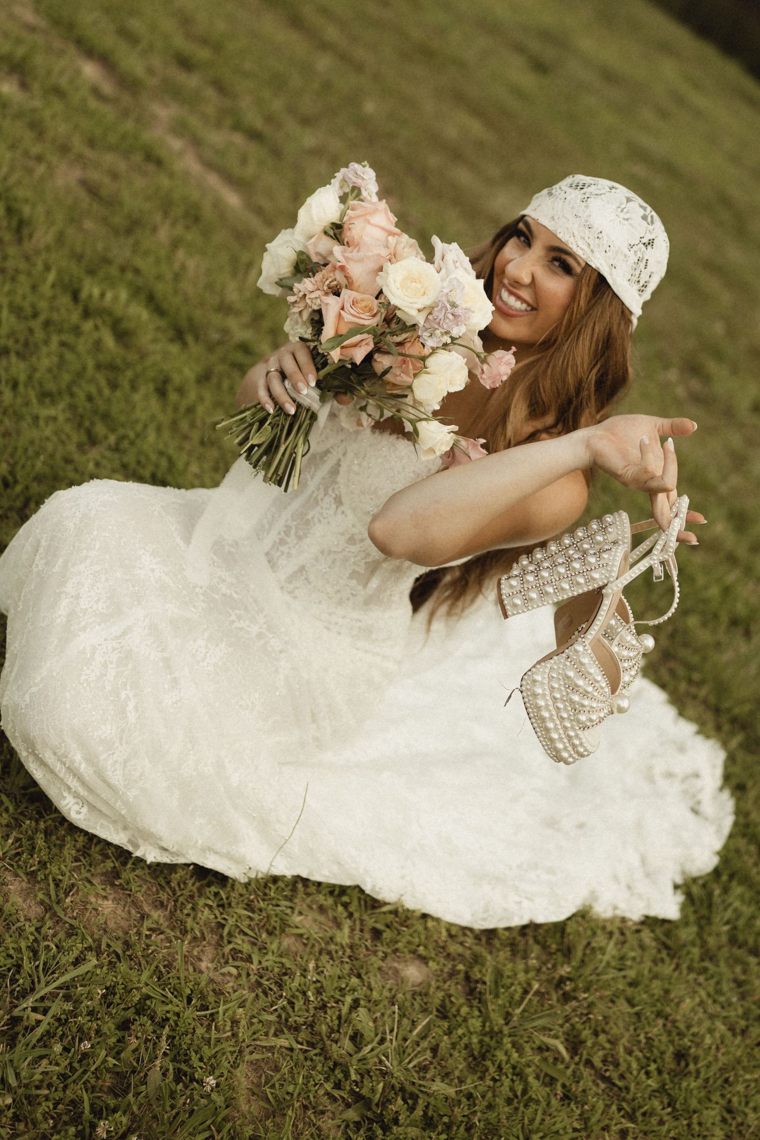 Smiling bride in white lace dress and cap sits on grass, holding floral bouquet and pearl heels outdoors.