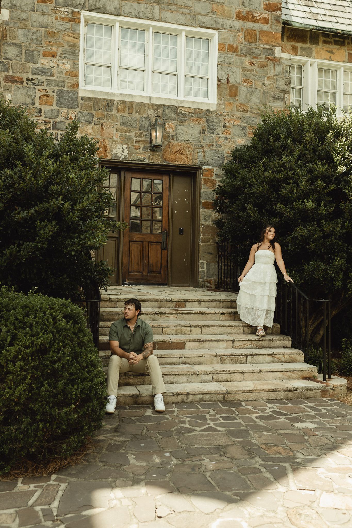 A couple poses on stone steps of a rustic building, man seated casually while woman in white dress descends gracefully.